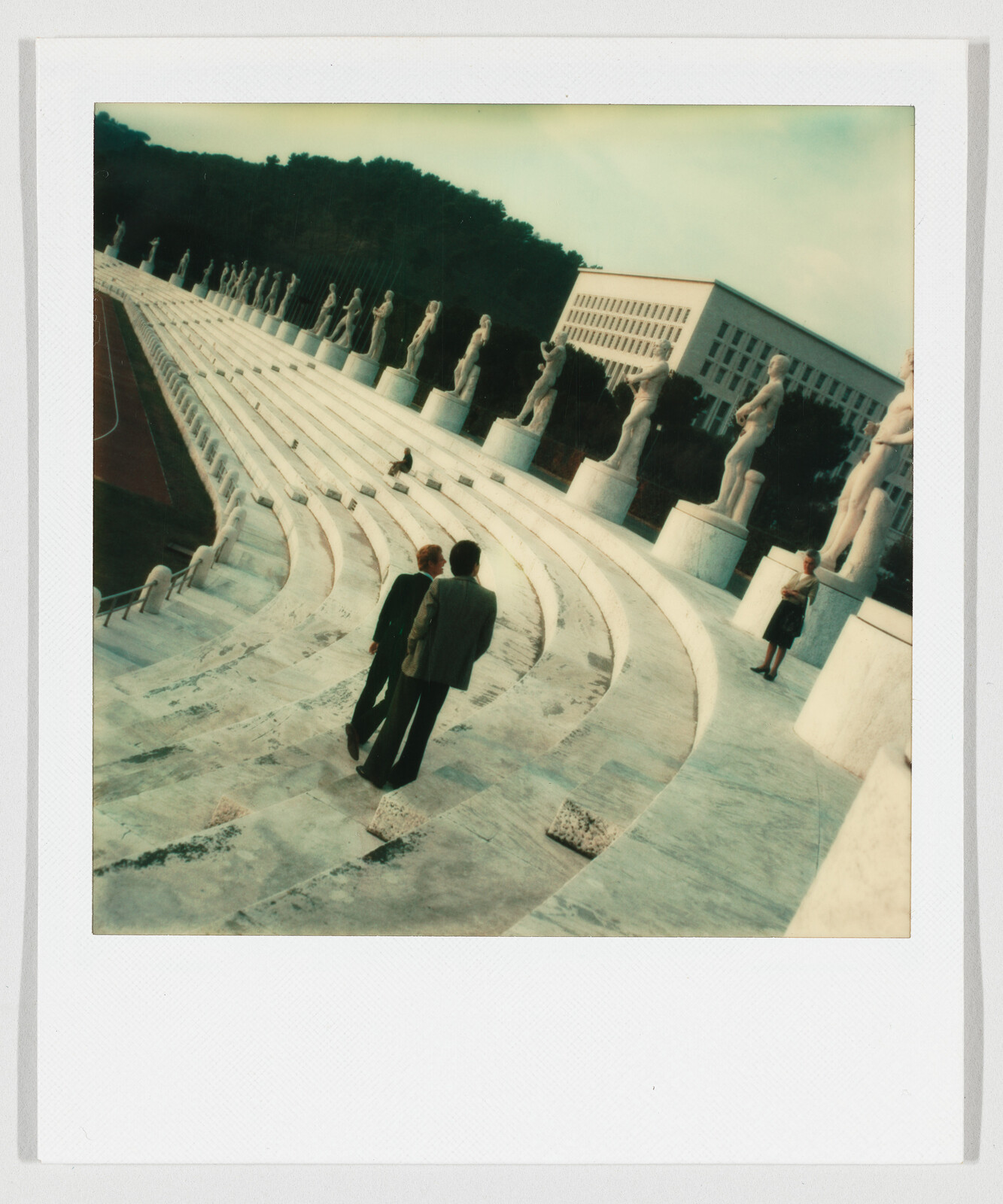 Two people walking down curved marble stadium steps lined with classical statues.