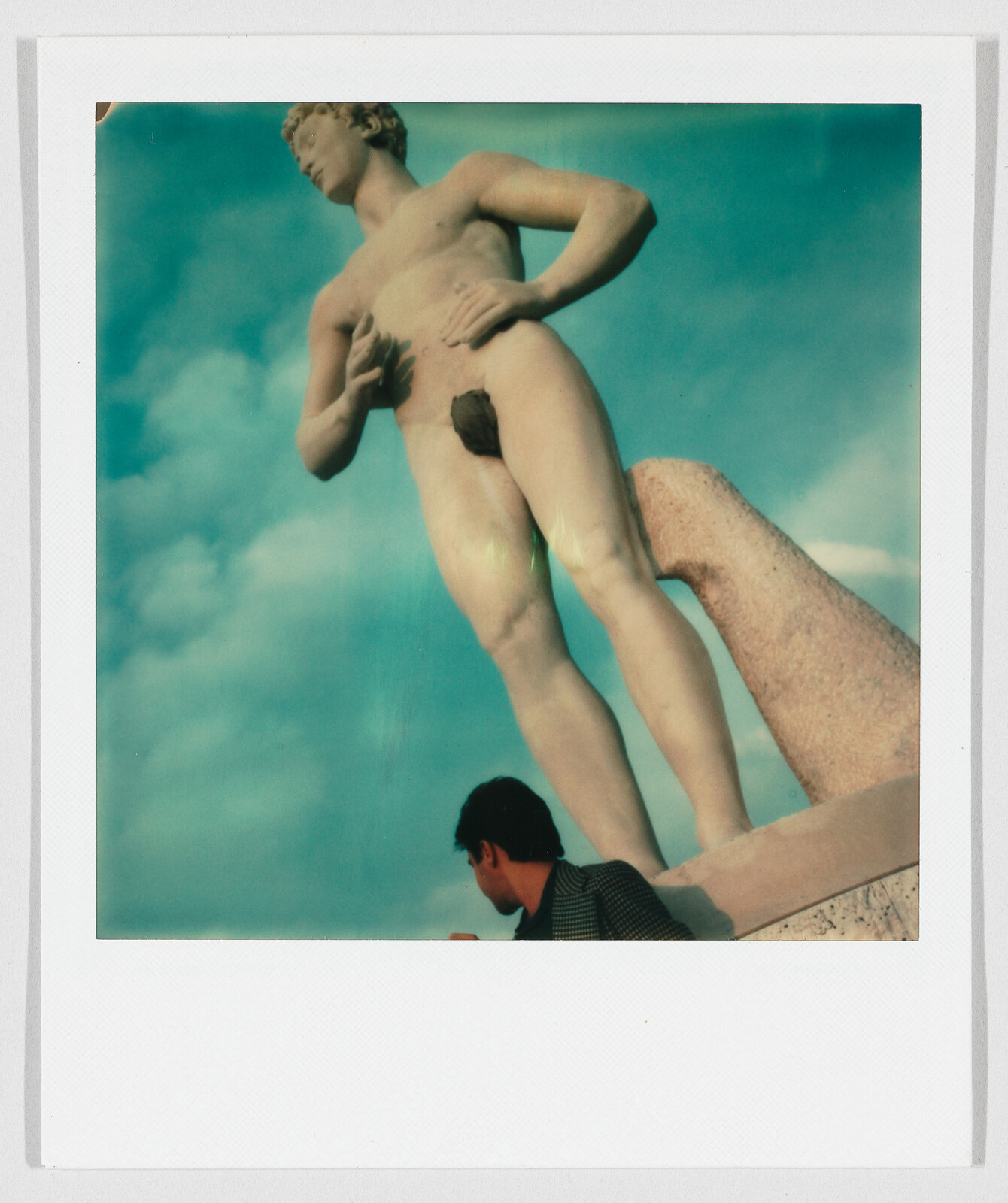 Large classical nude statue towering over a man looking up against a blue sky.
