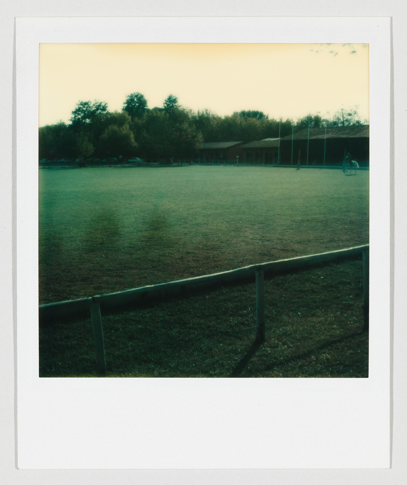 Empty grassy field with a wooden fence in front, trees and a building in the background.