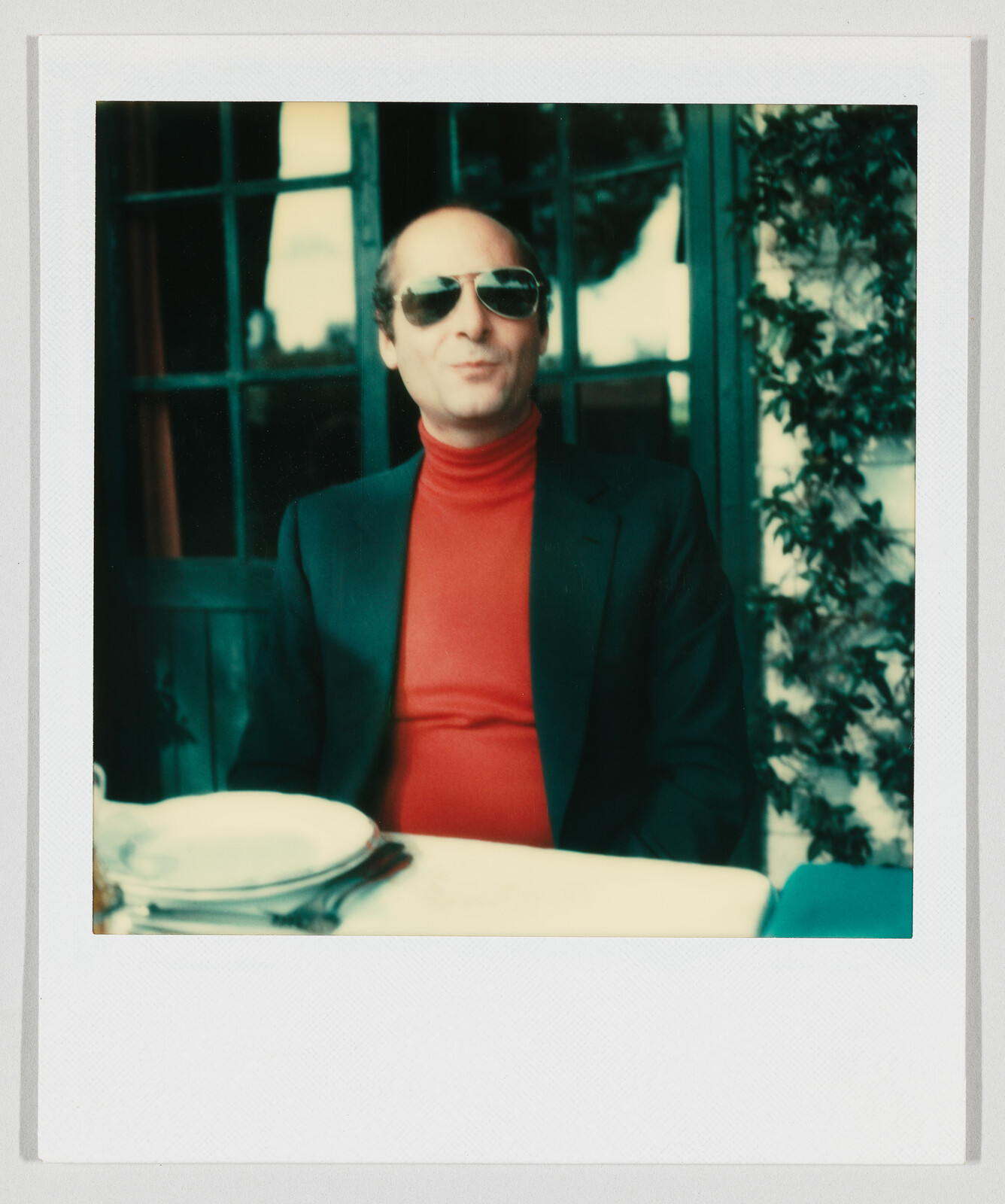 Man wearing sunglasses and a red turtleneck sits at an outdoor table with empty plates and utensils.