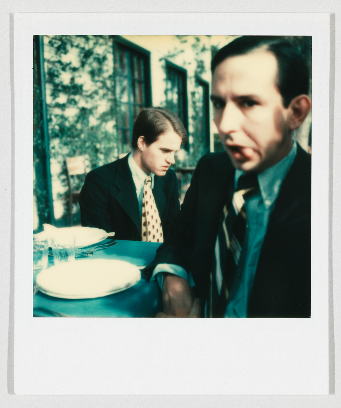 Two men in suits sit at a table while one looks down and the other stares toward the camera.