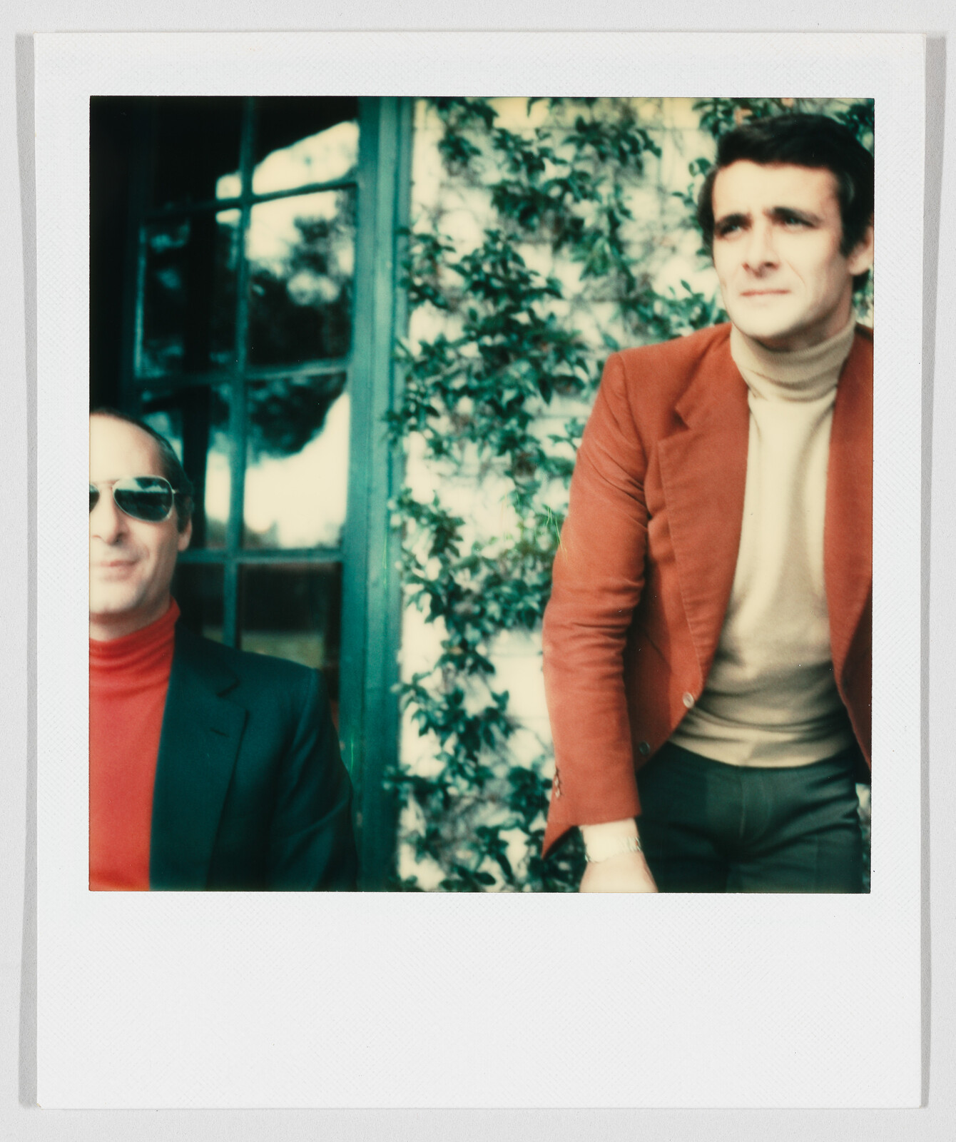 Two men pose outdoors, one standing in a rust blazer and the other seated wearing sunglasses.