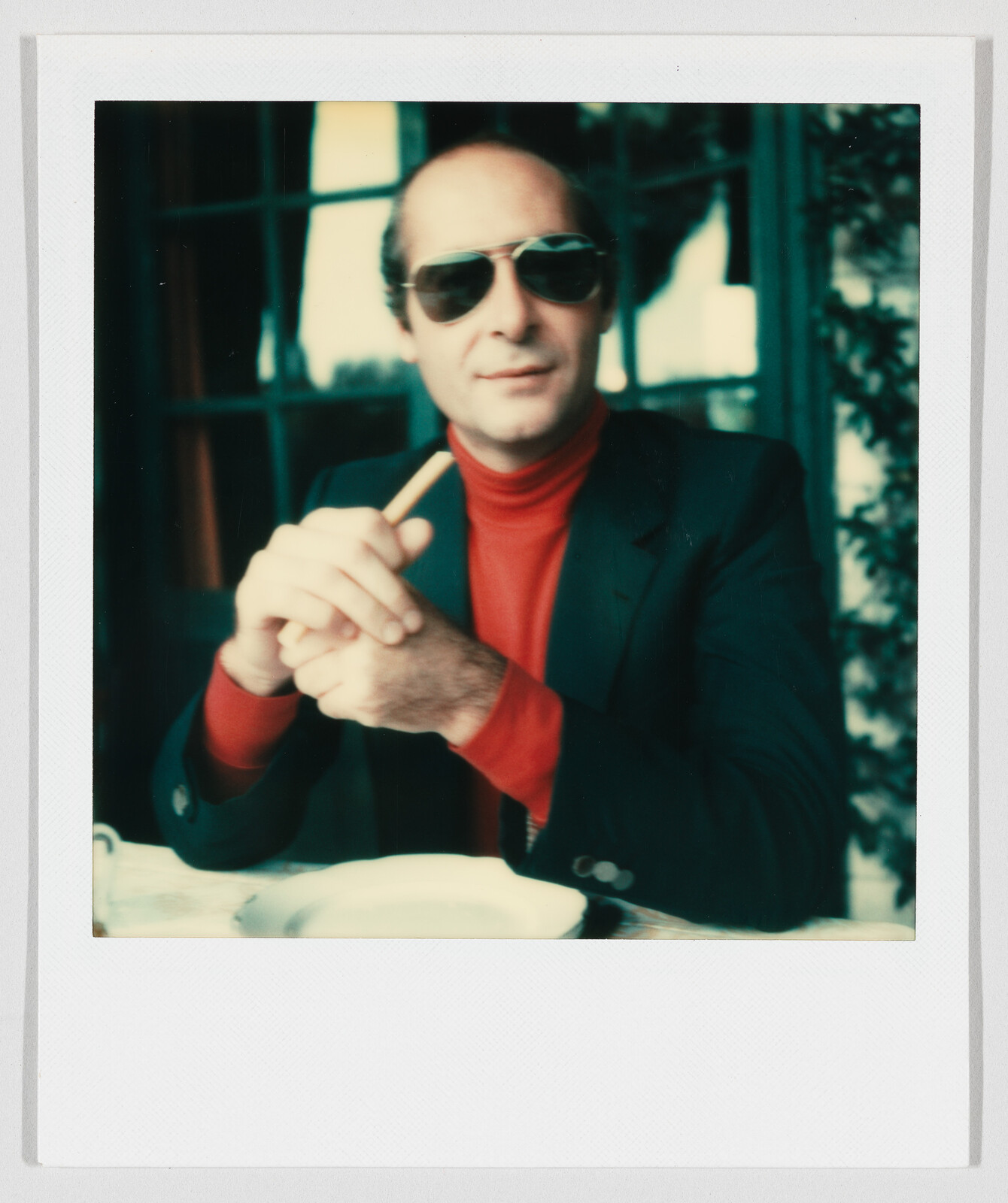 Man wearing sunglasses and red turtleneck holds a cigarette while sitting at a table.