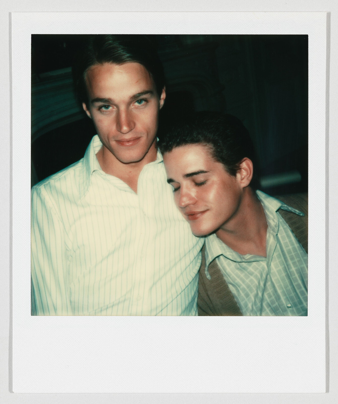 Two young men pose closely, one resting his head on the other's shoulder with eyes closed.