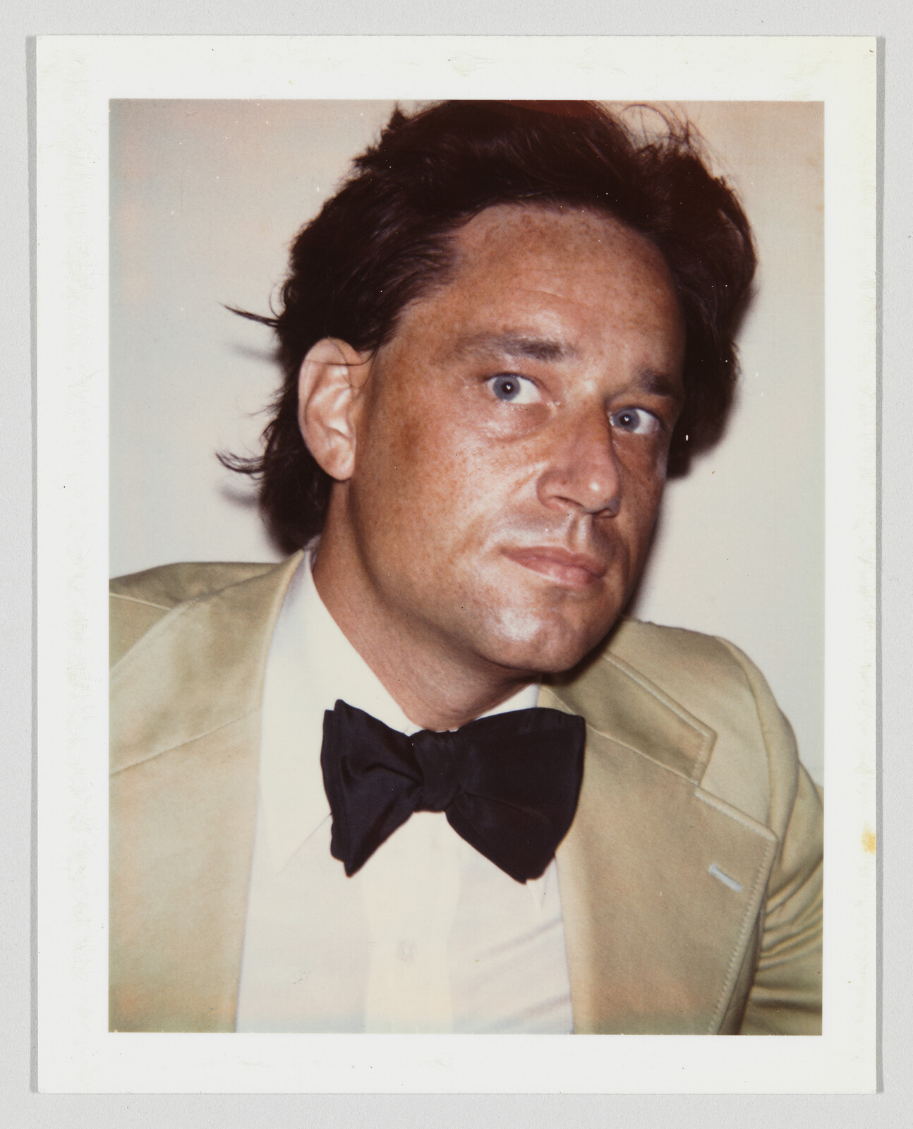 A man in a beige suit and black bow tie looks to the side with a serious expression.