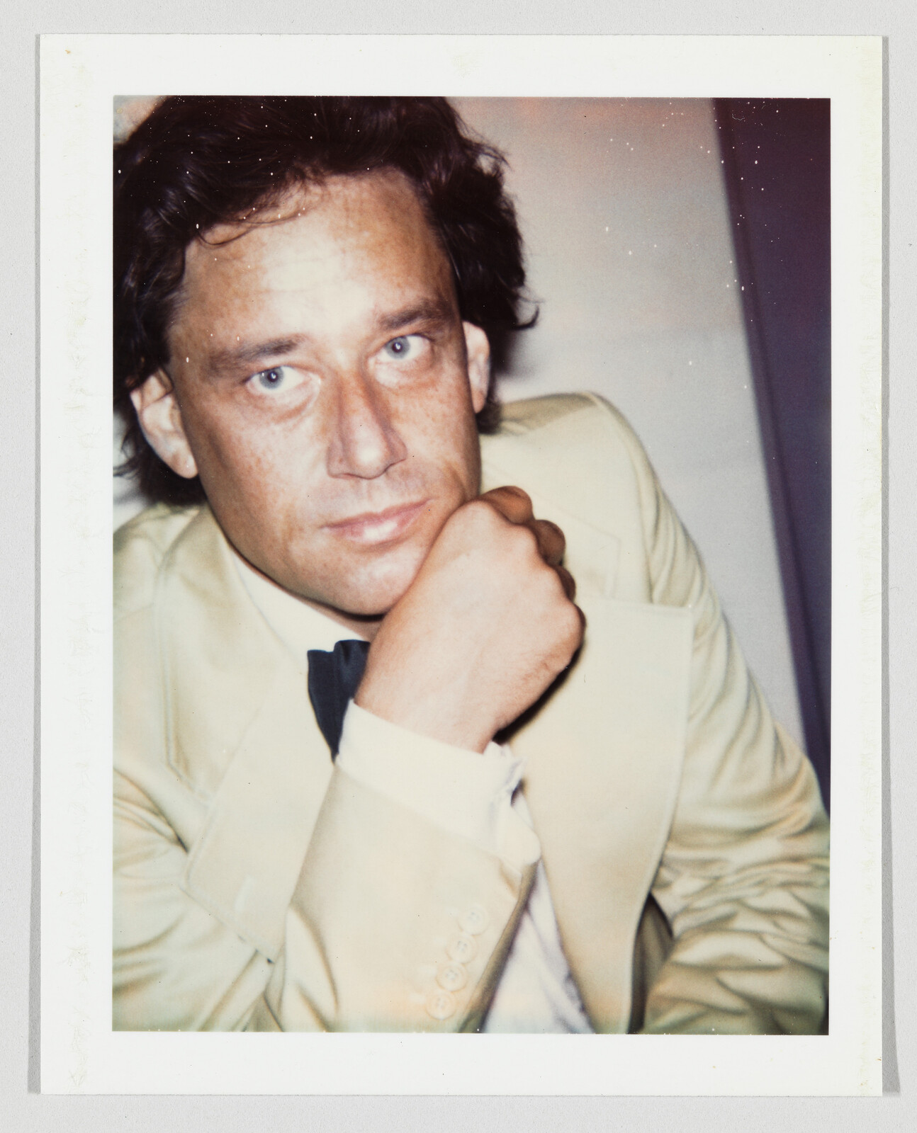 A man in a pale suit and bow tie rests his chin on his fist while looking at the camera.