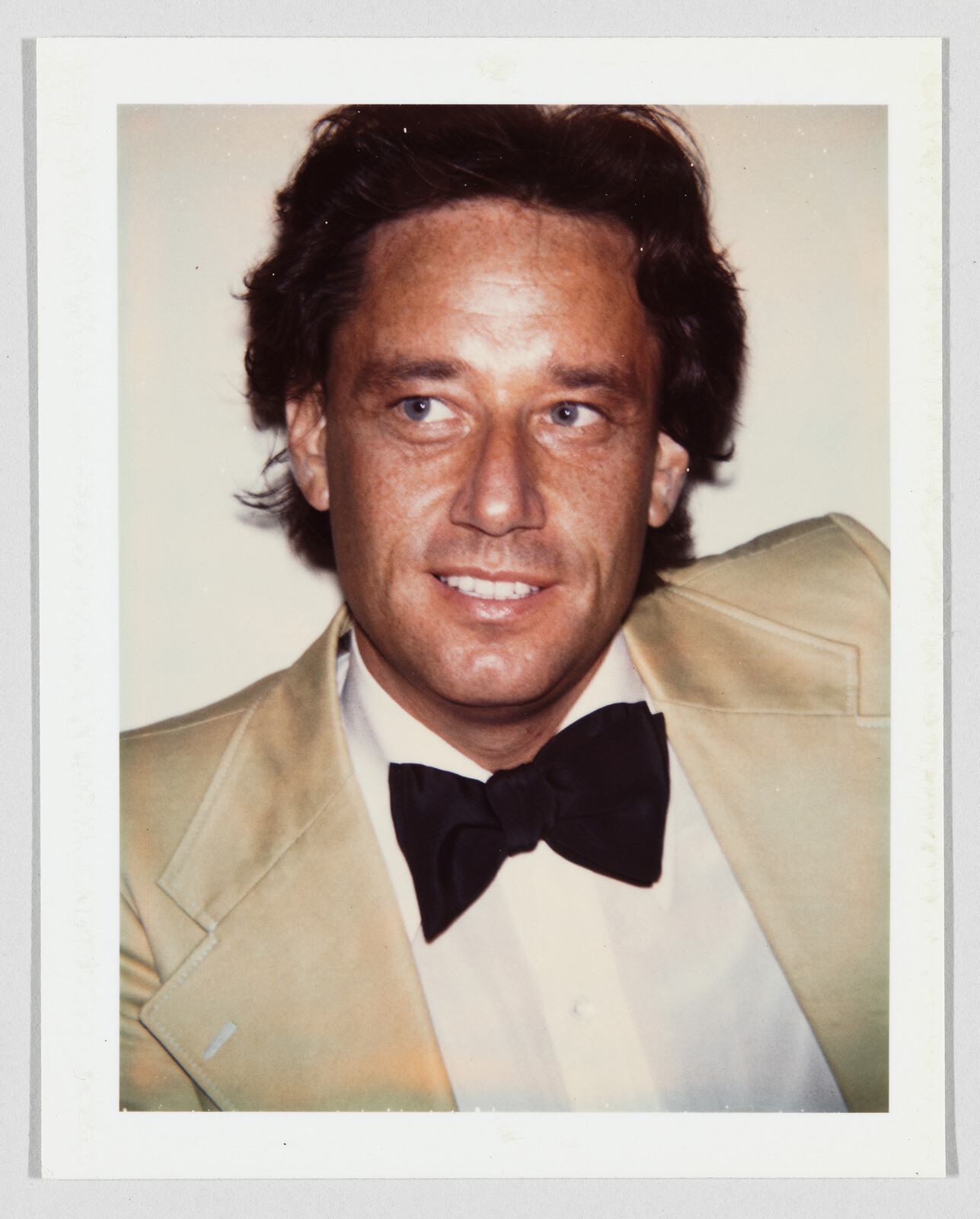 A man in a tan suit and black bow tie looks slightly to the side with a smile.