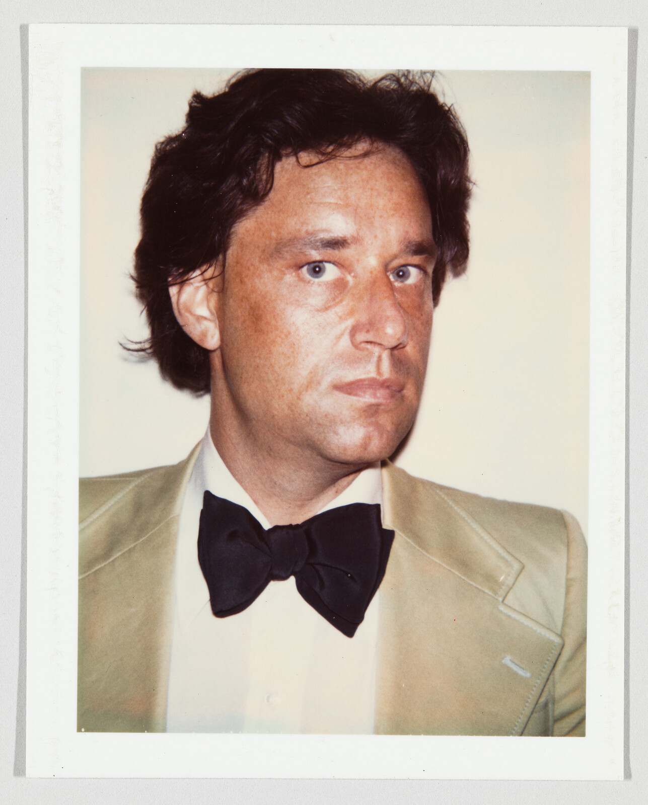 A man in a tan suit and black bow tie looks slightly to the side with a serious expression.