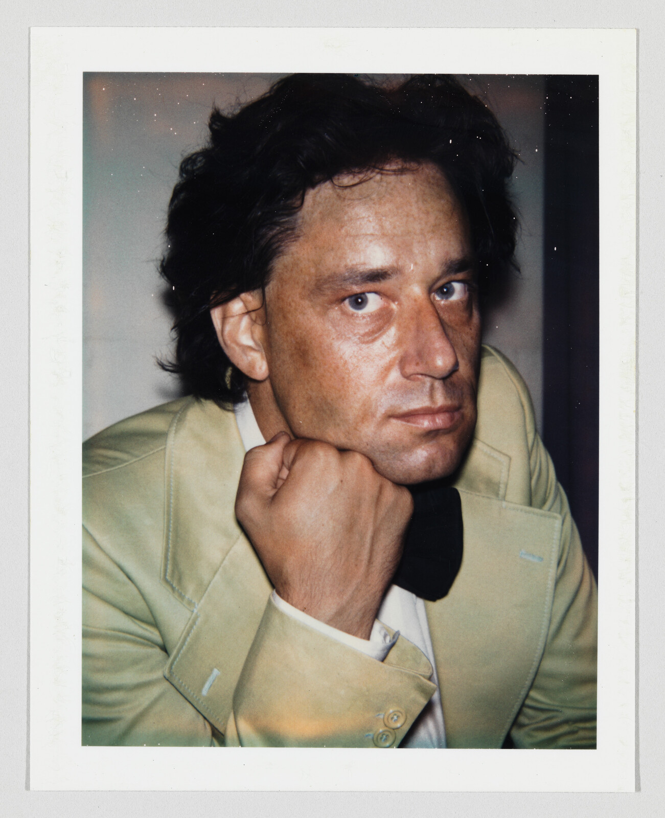 Man with dark hair wearing a light-colored jacket rests his chin on his hand, looking at the camera.