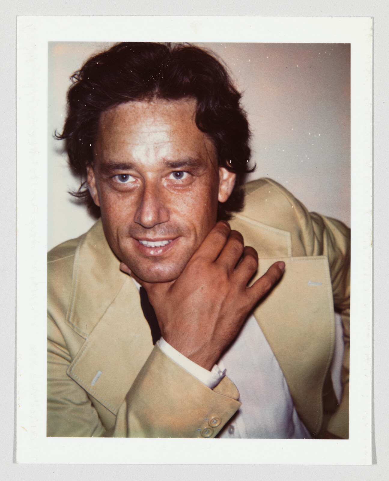 A smiling man in a beige suit rests his hand on his neck while looking forward.