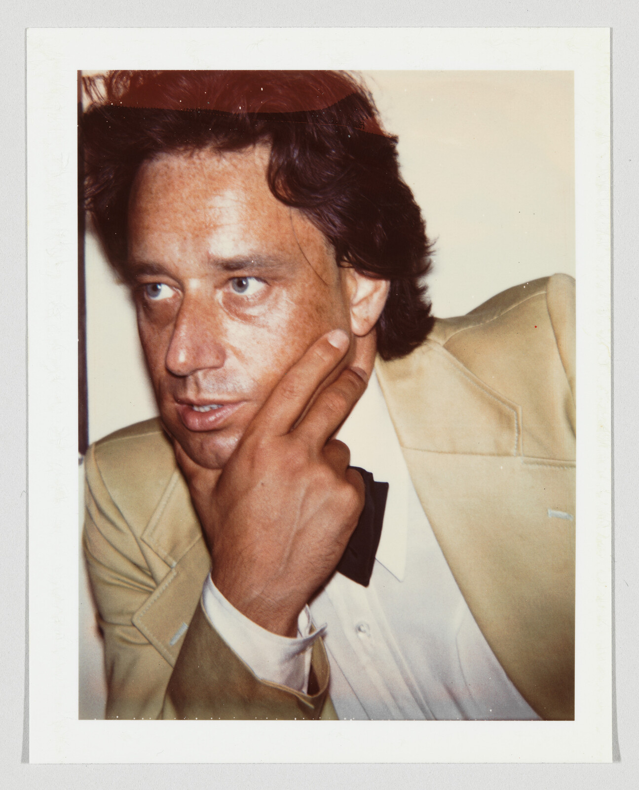 A man in a tan suit and bow tie rests his hand on his chin while looking sideways.