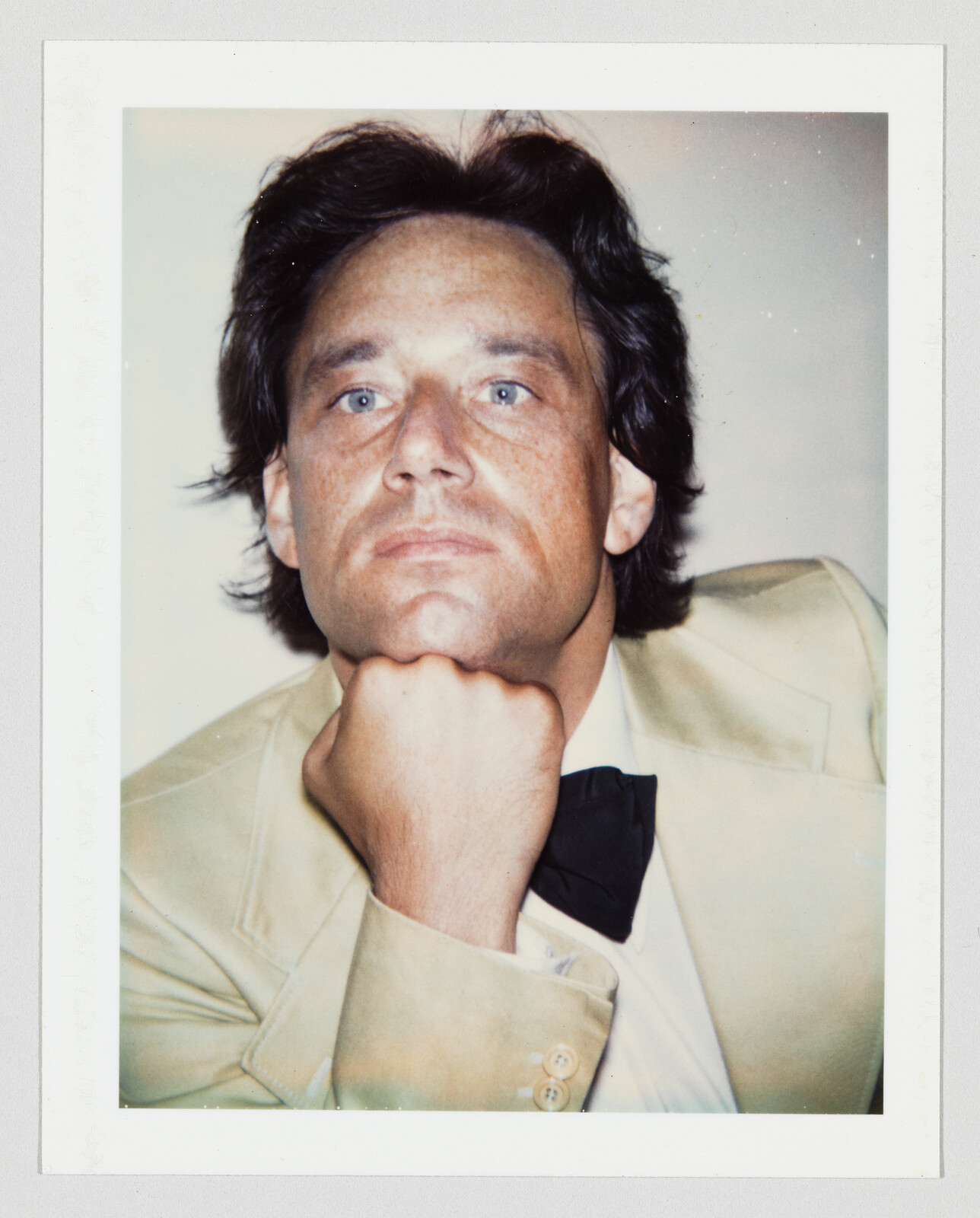 Man with medium-length dark hair wearing a tan suit and black bow tie, resting chin on his hand.