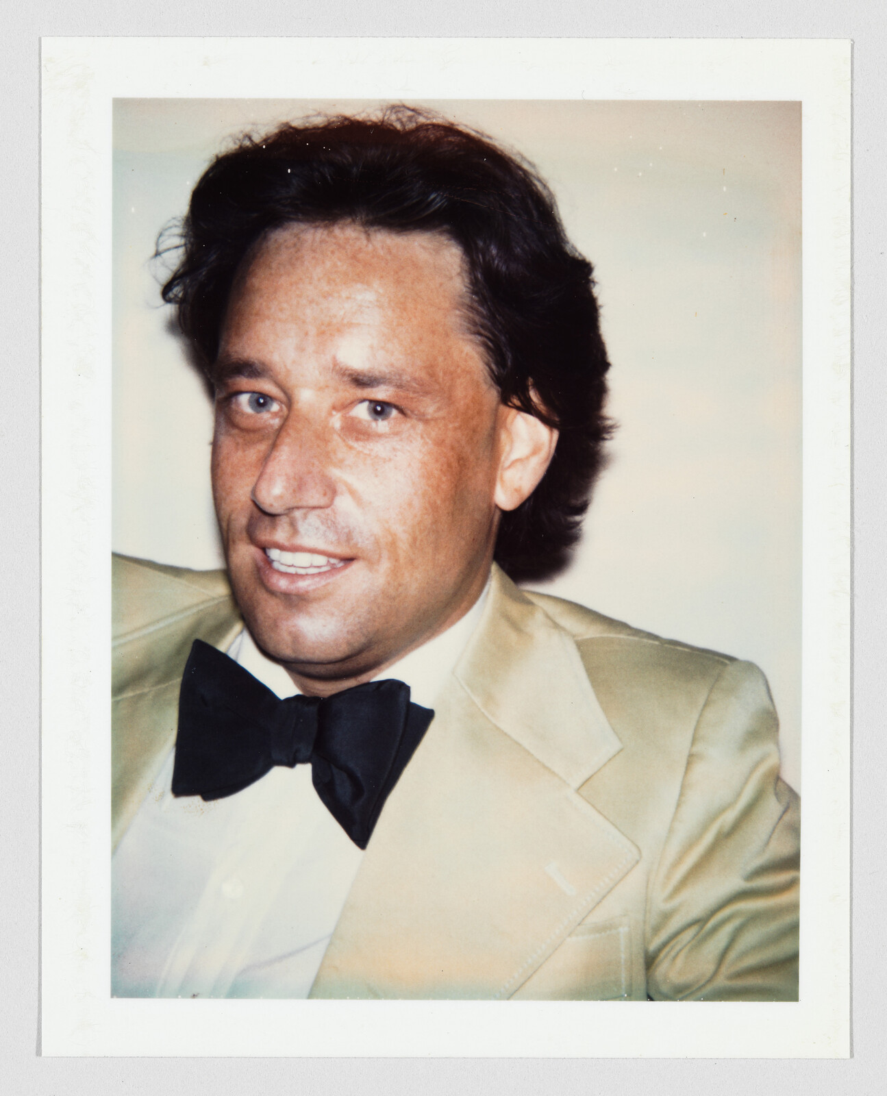 Man wearing a beige tuxedo jacket and black bow tie smiling toward the camera.