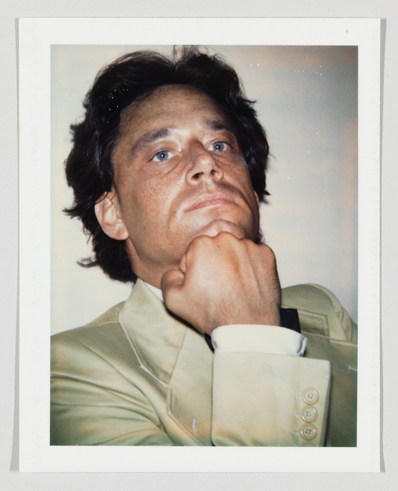 A man in a beige suit rests his chin on his fist while looking upward thoughtfully.