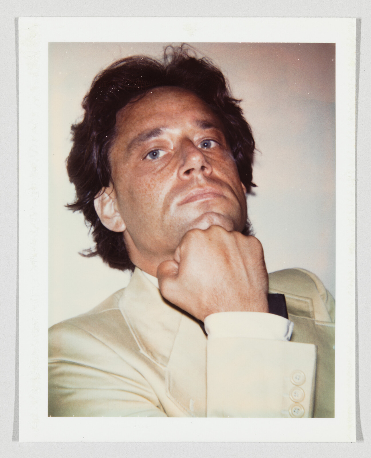 A man in a light suit rests his chin on his fist while looking upward confidently.