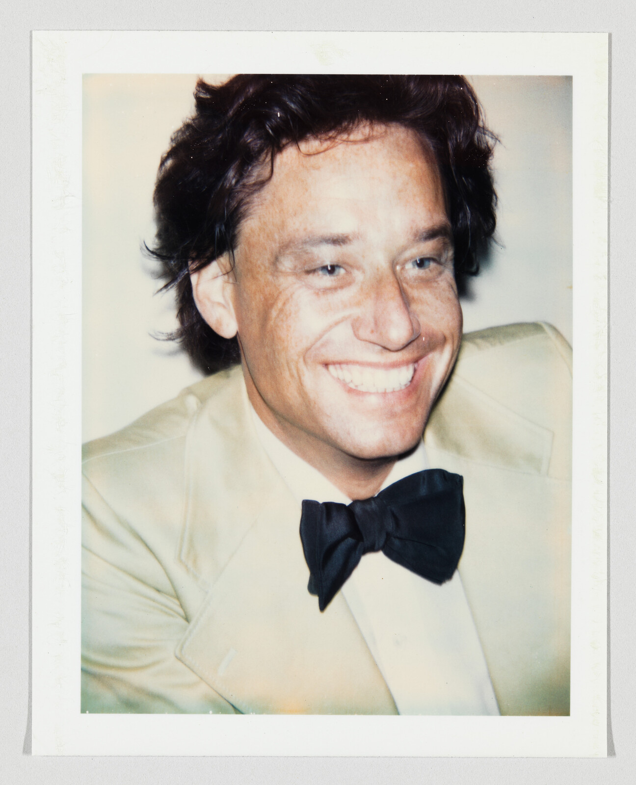 Smiling man in a light tuxedo jacket wearing a black bow tie in a close-up portrait.