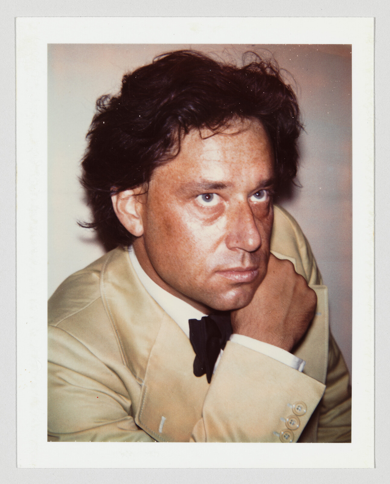 Man in a tan suit and black bow tie resting his chin on his hand, looking upward.