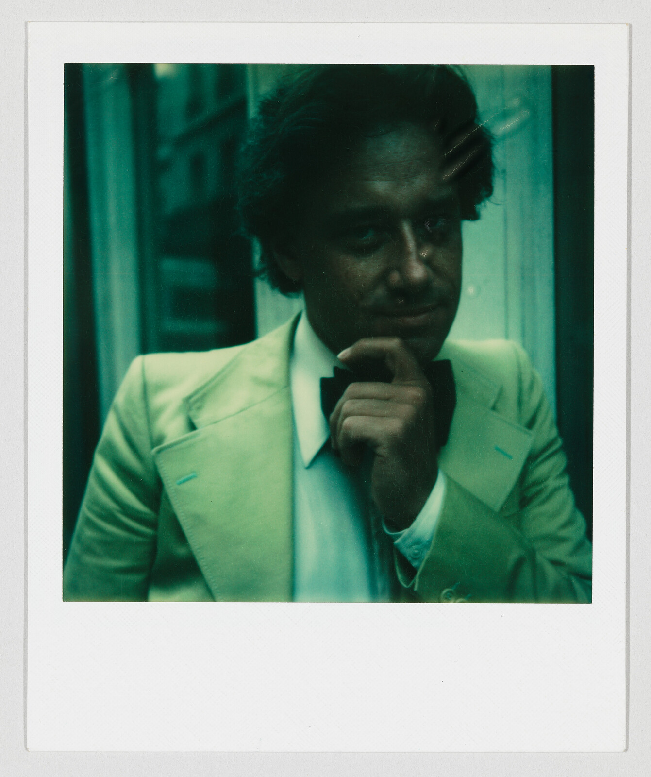 Man in a light-colored suit and bow tie stands indoors, resting his chin on his hand.