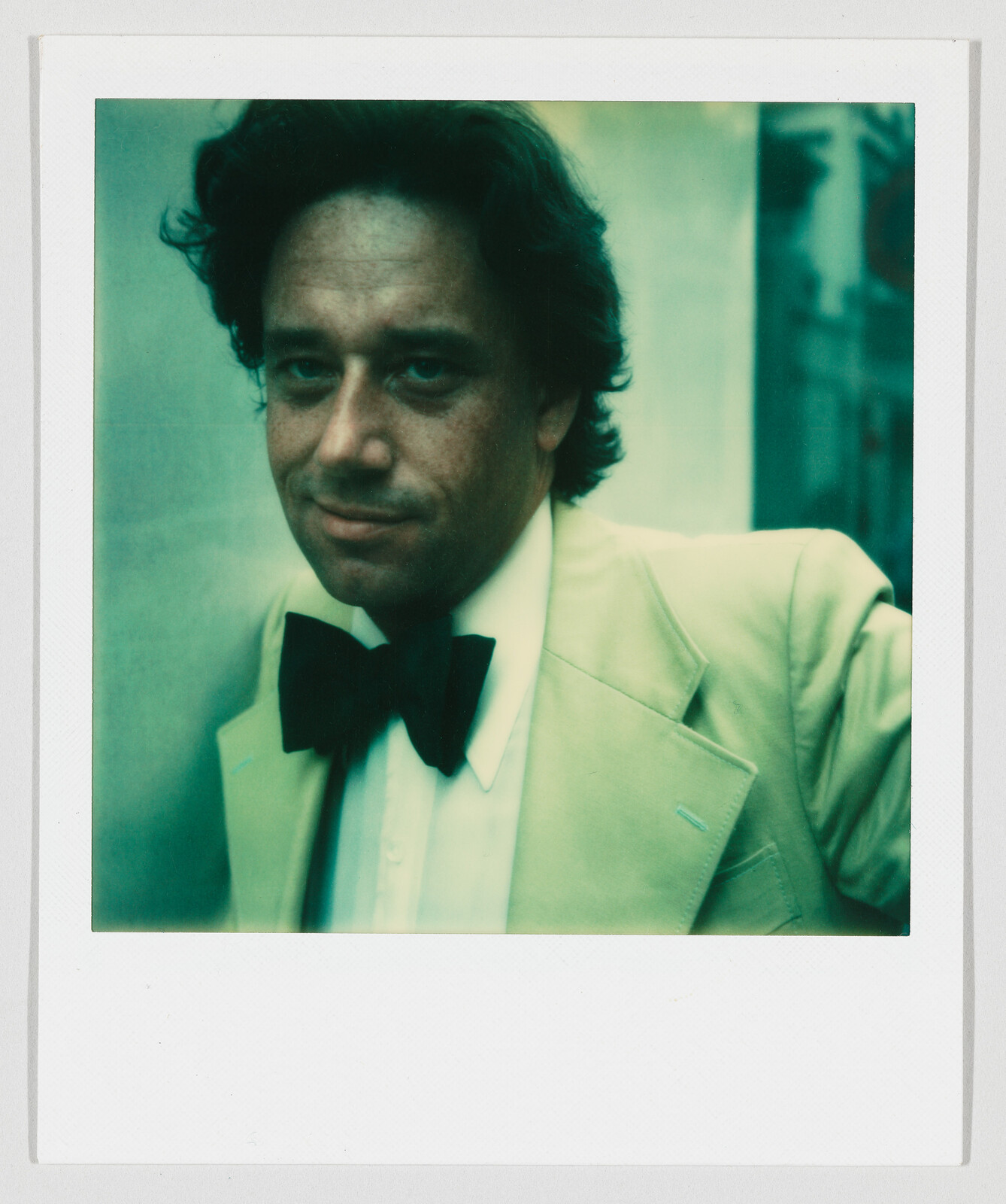 A man wearing a light-colored suit and black bow tie looks toward the camera with a slight smile.