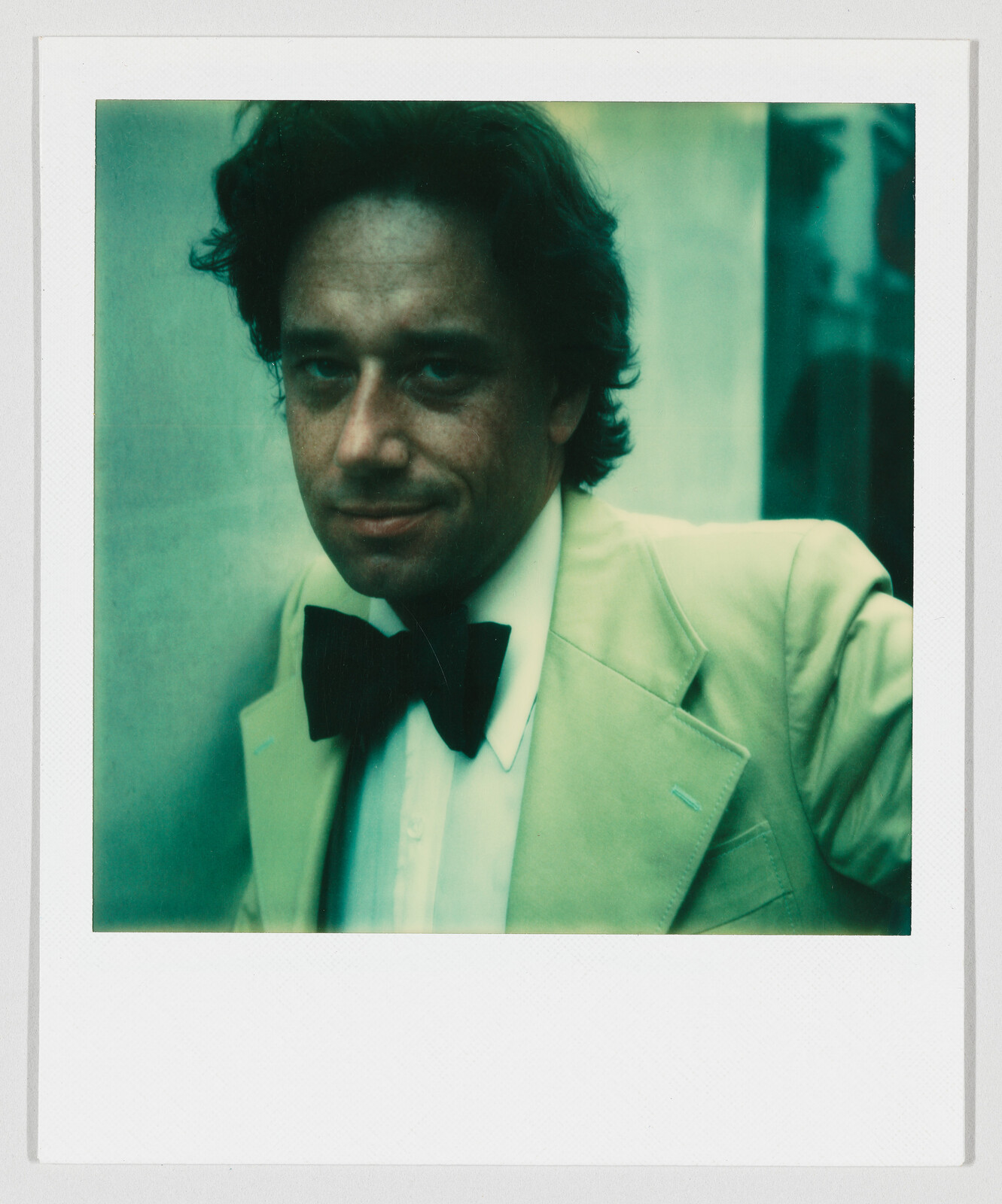 A man wearing a pale suit jacket and black bow tie looks toward the camera with a slight smile.