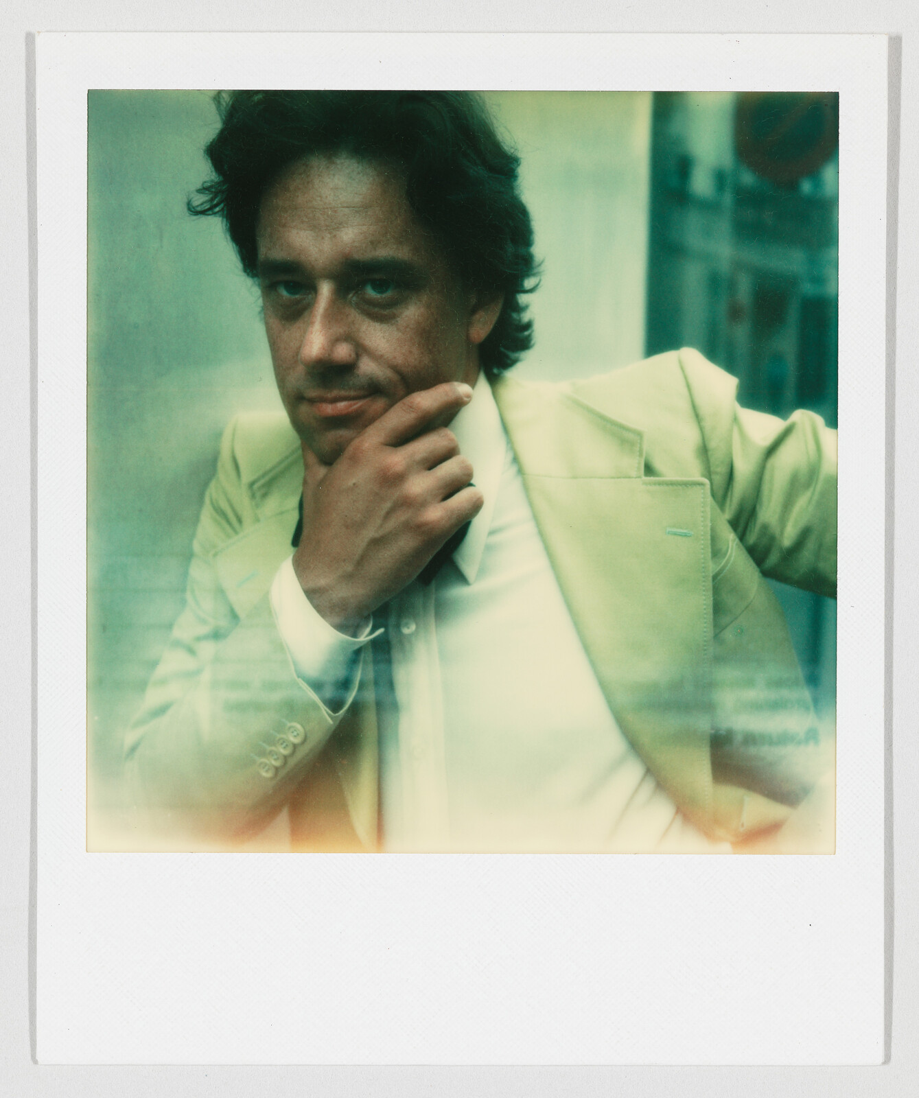 A man in a light suit leans with one arm and touches his chin in a thoughtful pose.