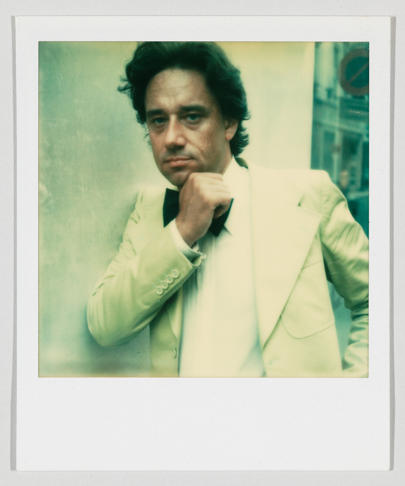 Man in a white tuxedo adjusts his bow tie while leaning against a wall, looking at camera.