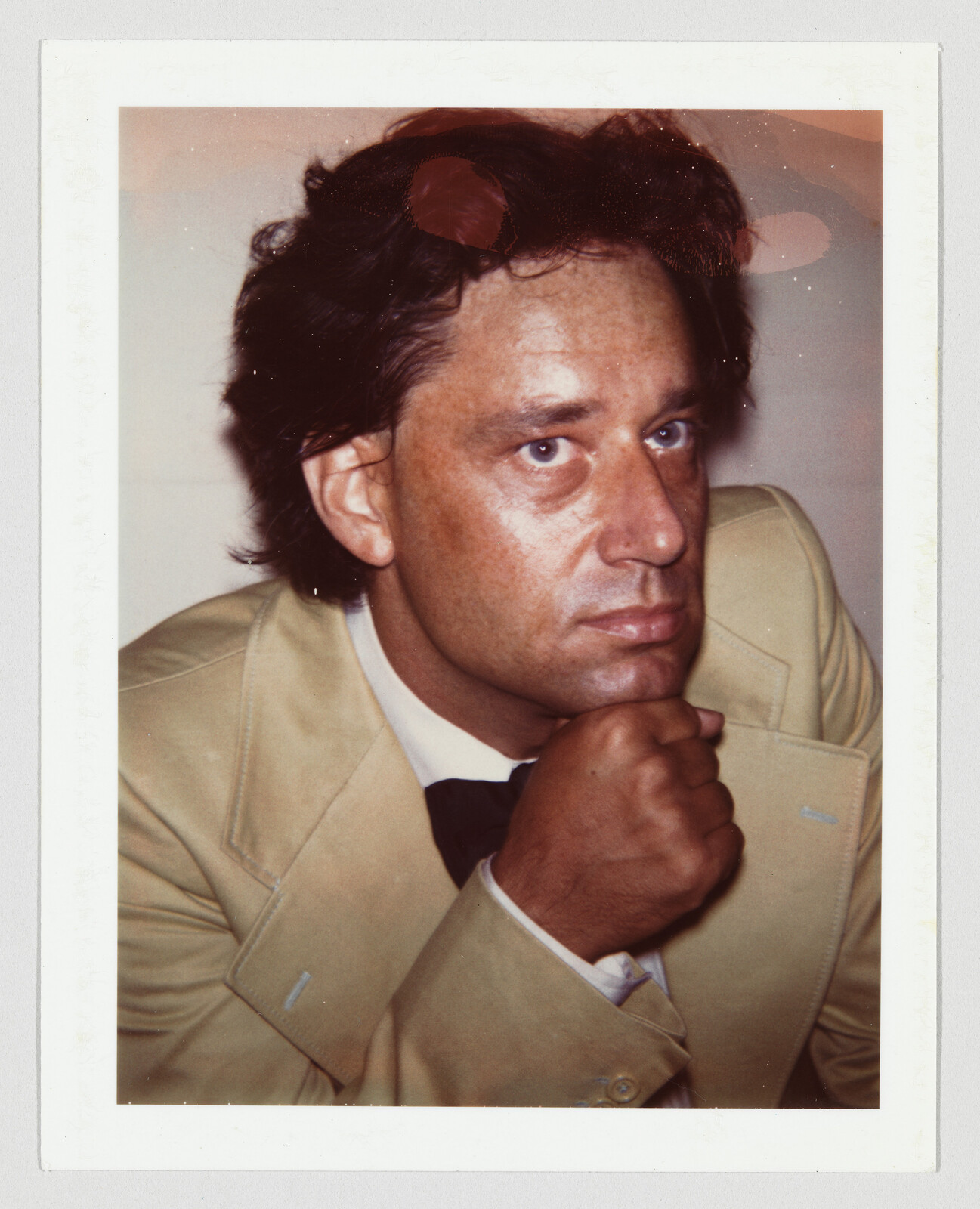 A man in a beige suit rests his chin on his hand while looking thoughtfully to the side.