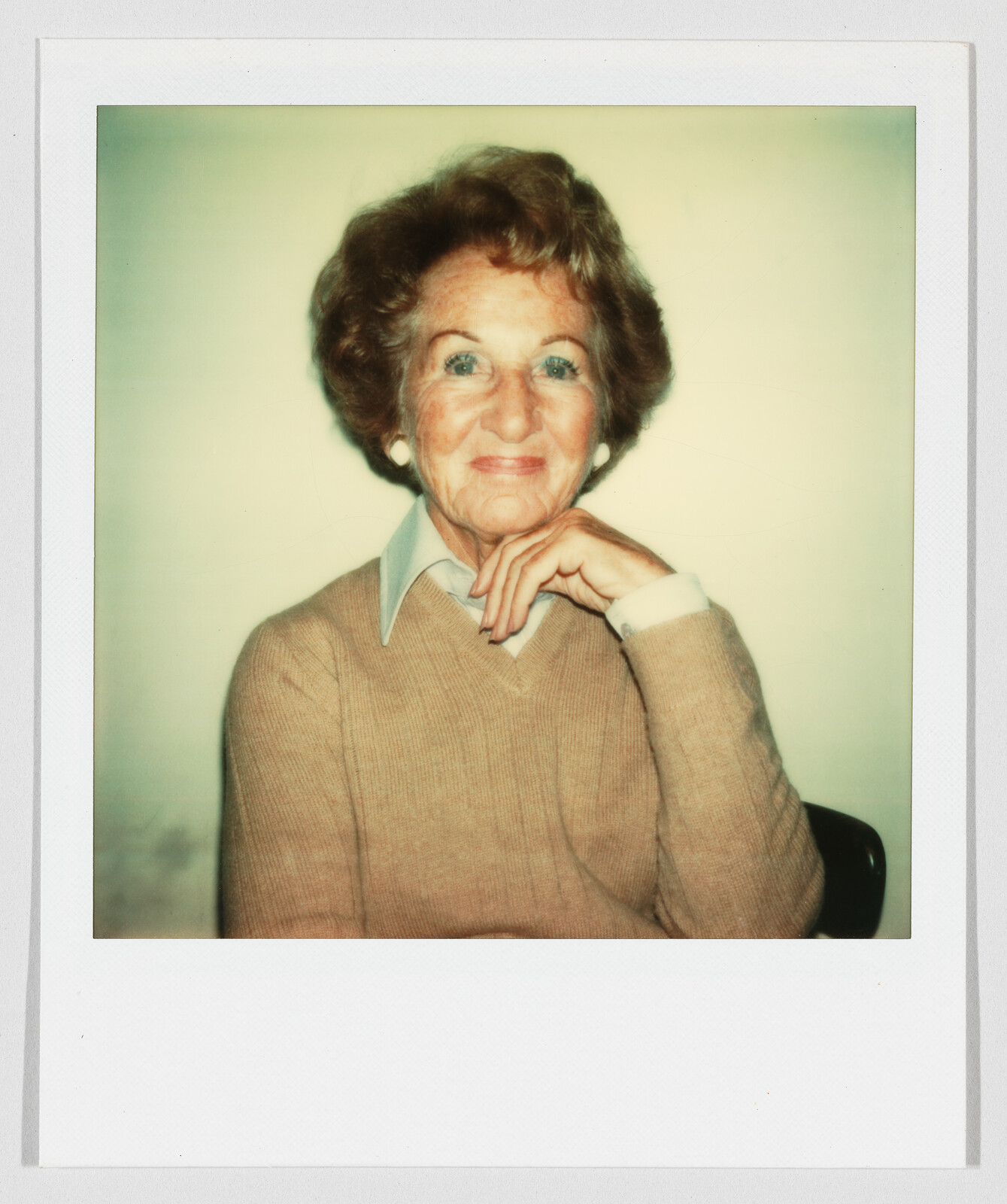 An older woman smiles gently with her hand resting under her chin, wearing a sweater.
