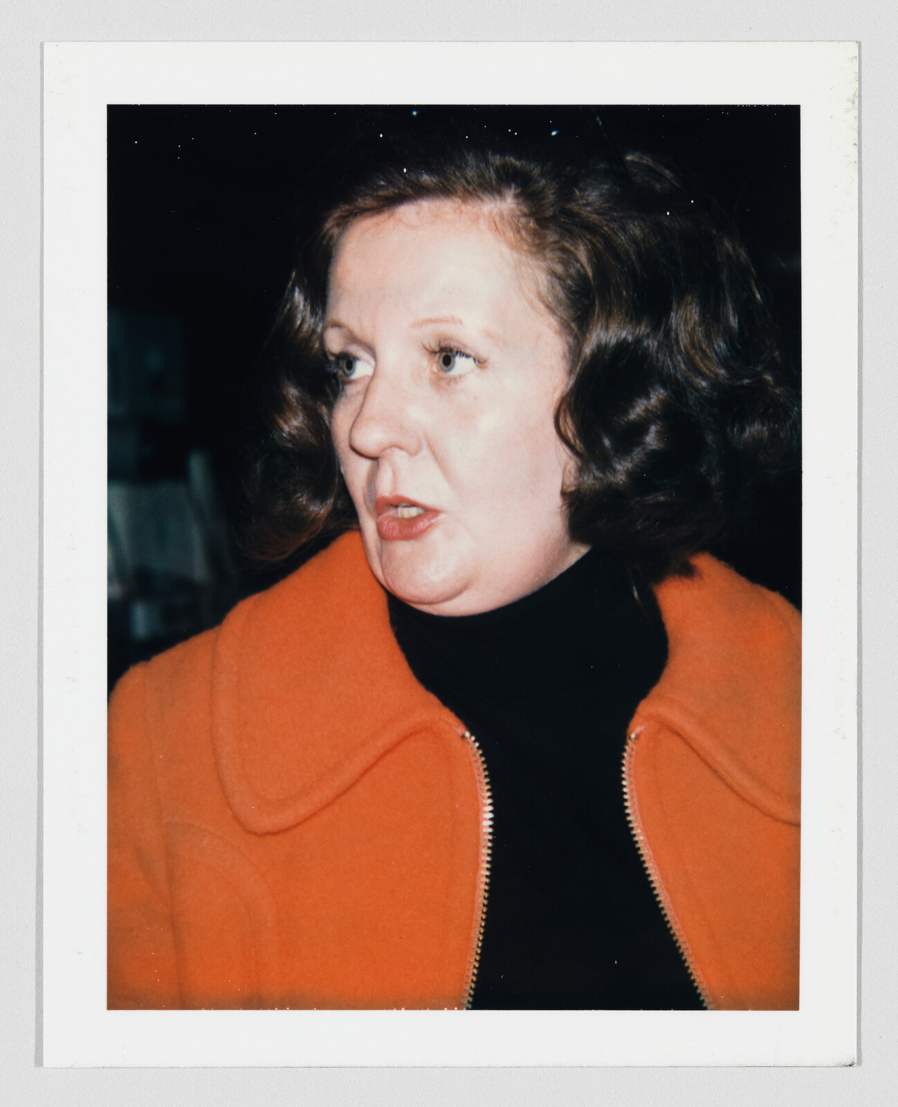 Middle-aged woman in an orange coat looking to the left while speaking.
