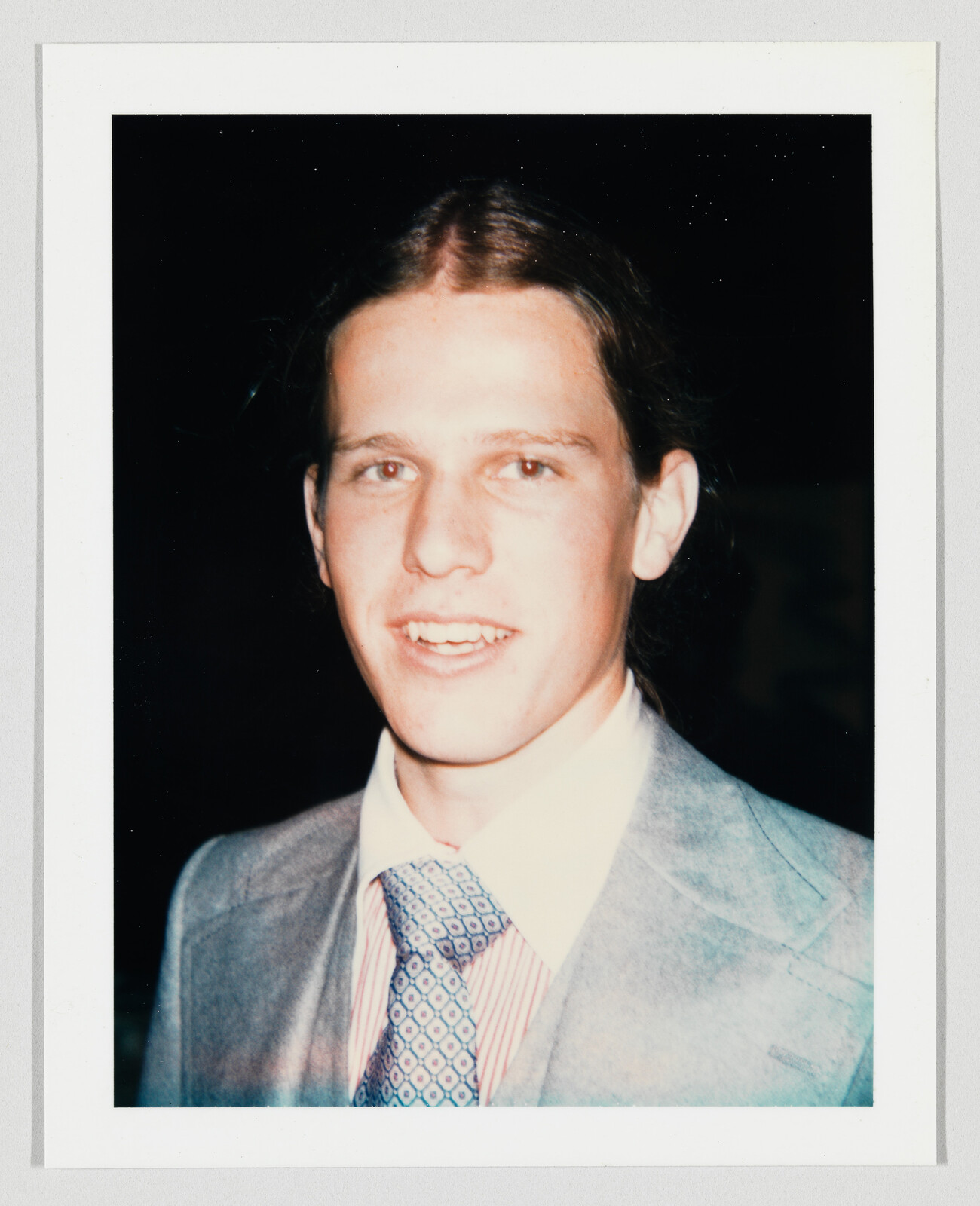 Young man smiling at camera while wearing a light gray suit, striped shirt, and patterned tie.