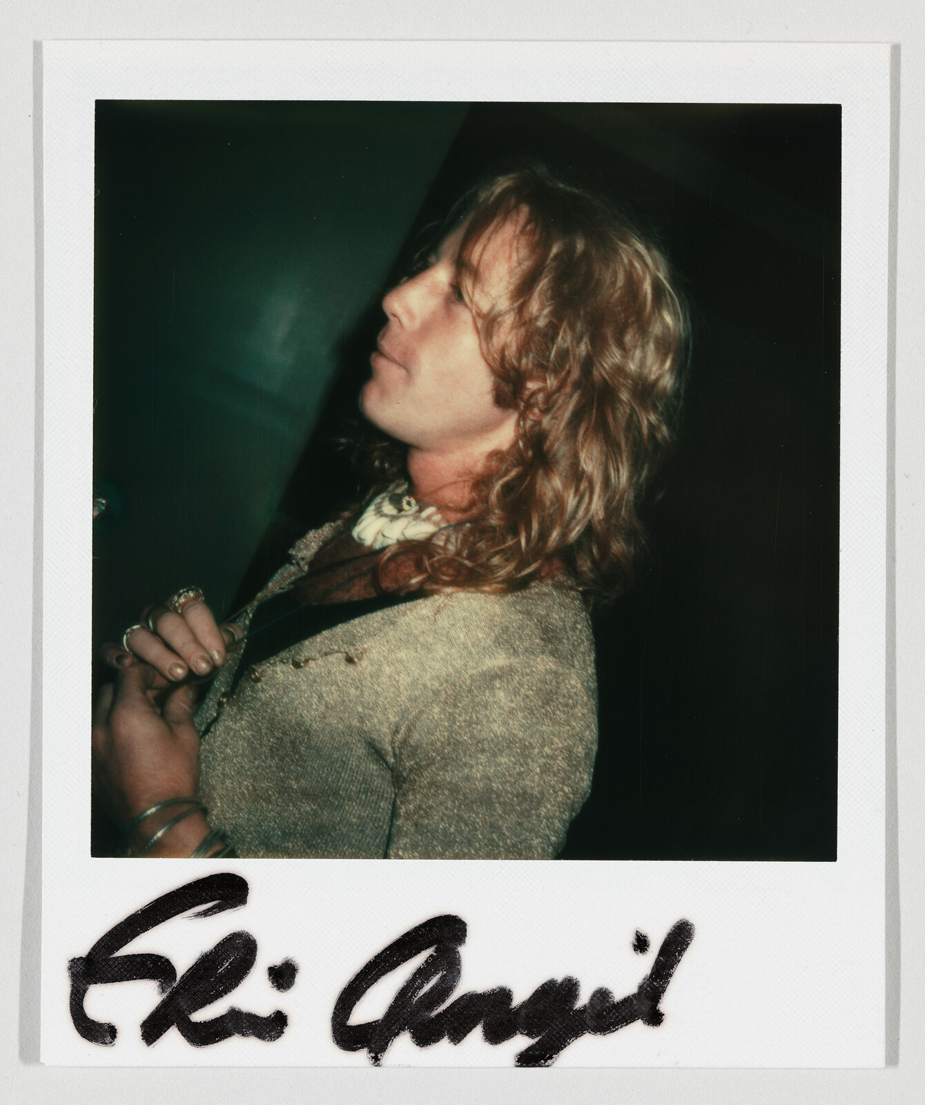 Person with long curly hair wearing a sweater and scarf, holding a strap. Handwritten signature at the bottom.