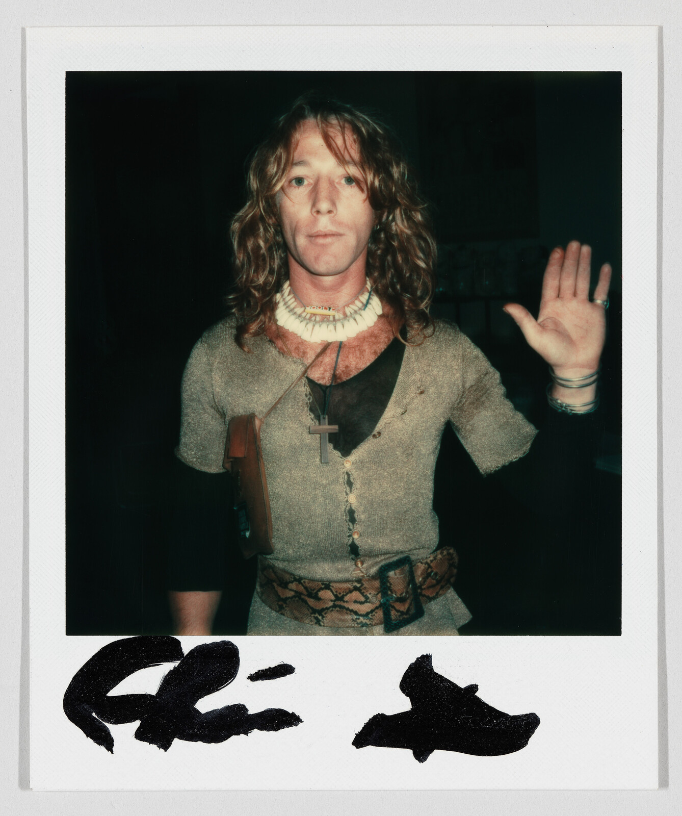 Man with long hair wearing necklaces and a belt raises his right hand.