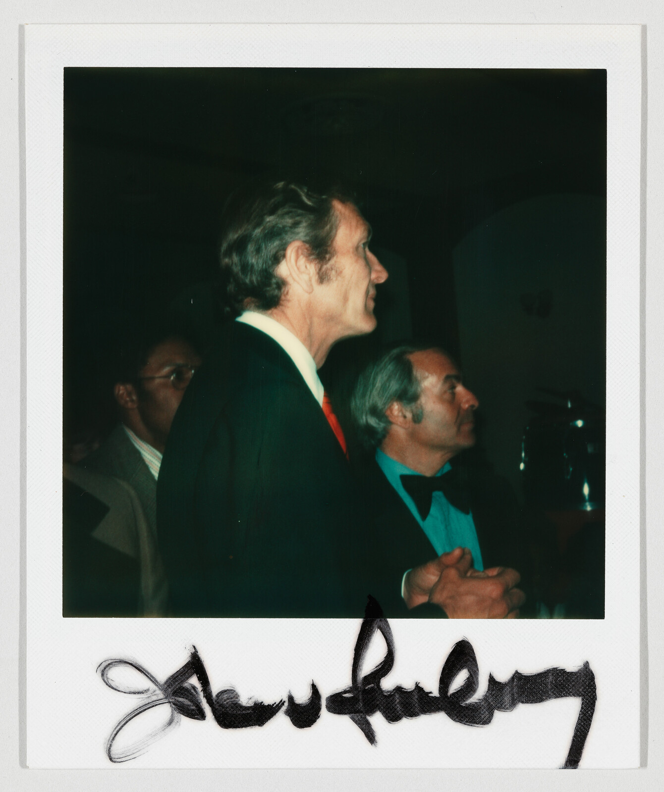Tall man in suit and red tie stands attentively next to a man in a bow tie.