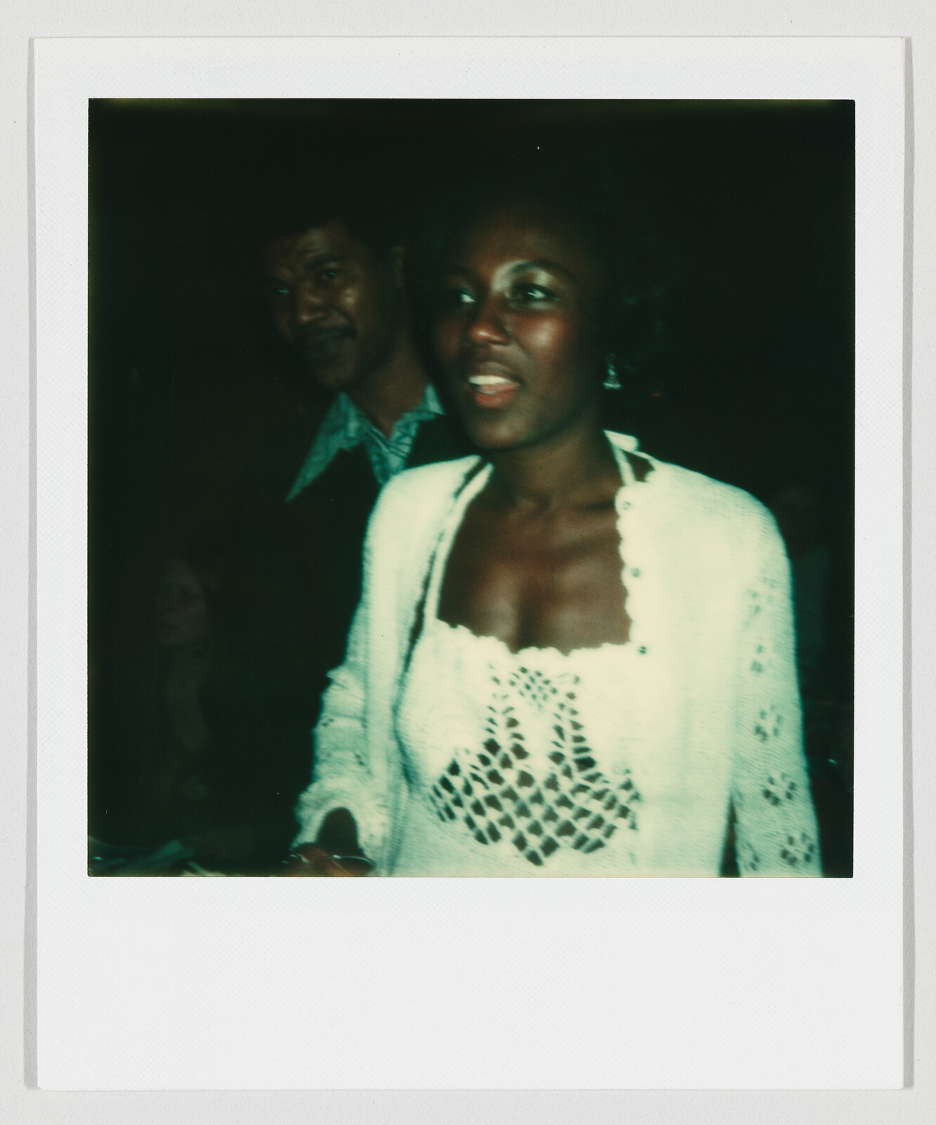Woman in a white crocheted top speaking while a man stands behind her in dim lighting.