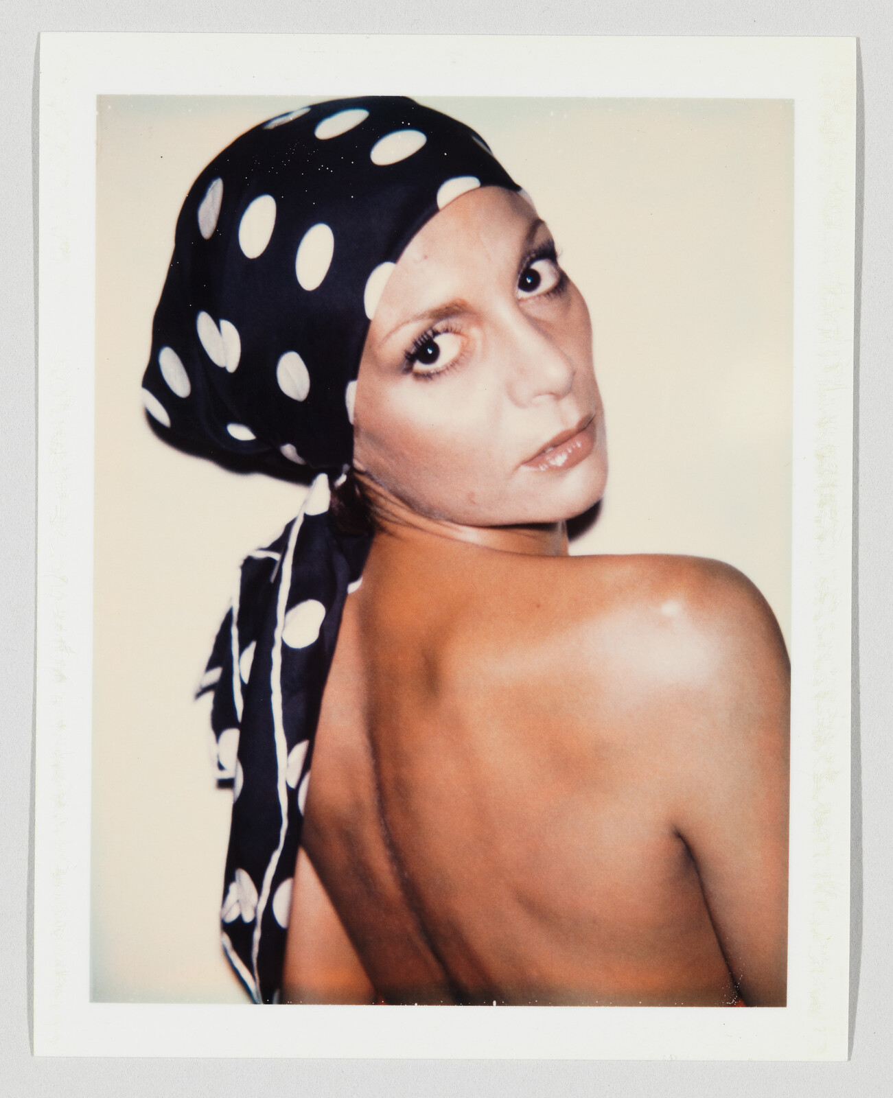 Woman wearing a black polka-dot headscarf looks over her shoulder, showing her bare back.