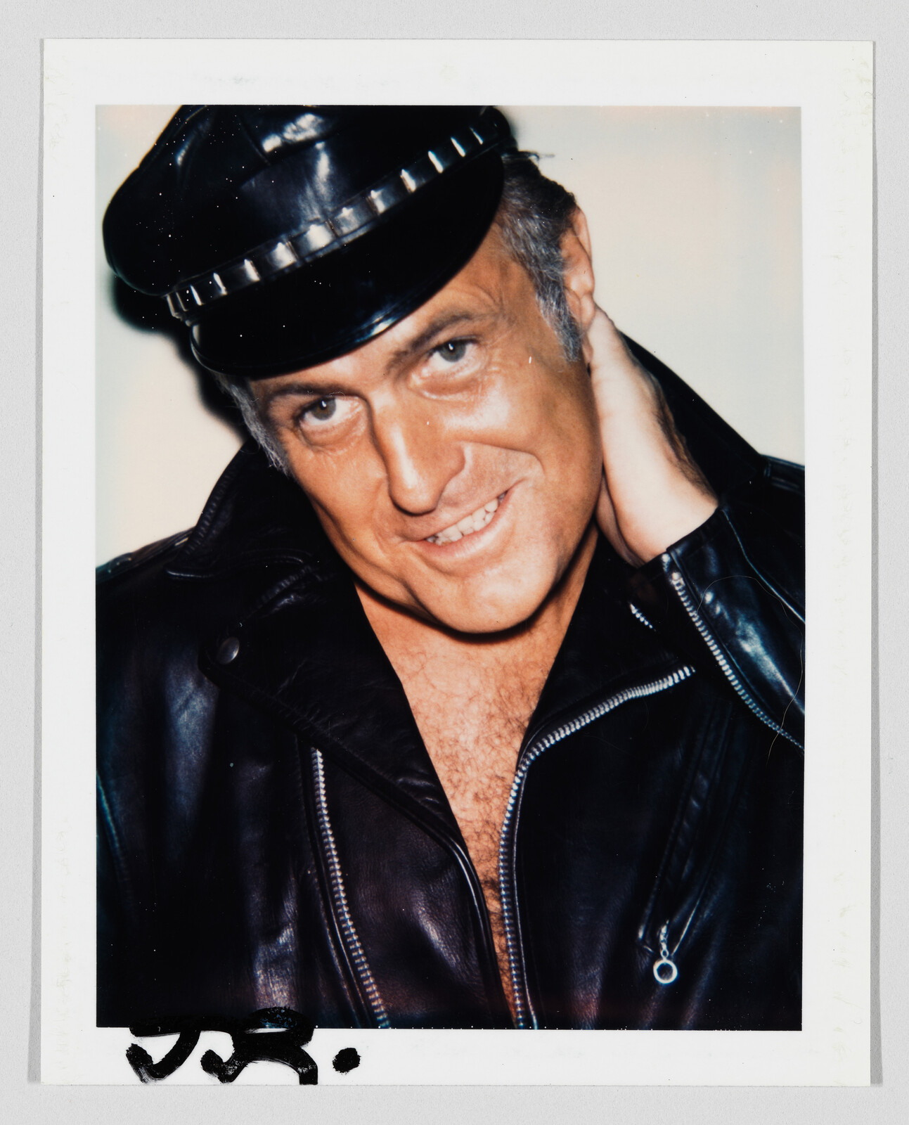 Man in black leather cap and jacket smiles while touching the back of his neck.