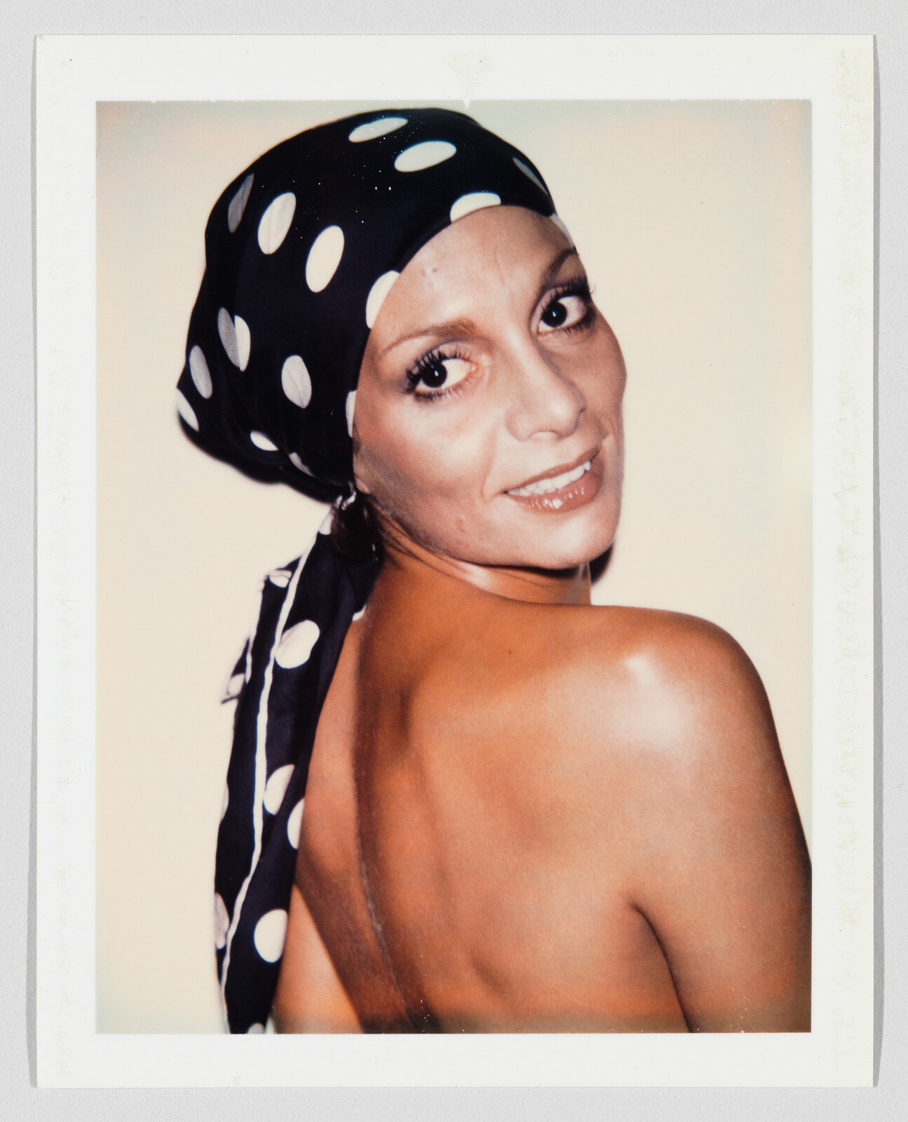 A woman wearing a black polka-dot headscarf looks over her shoulder with a smile.