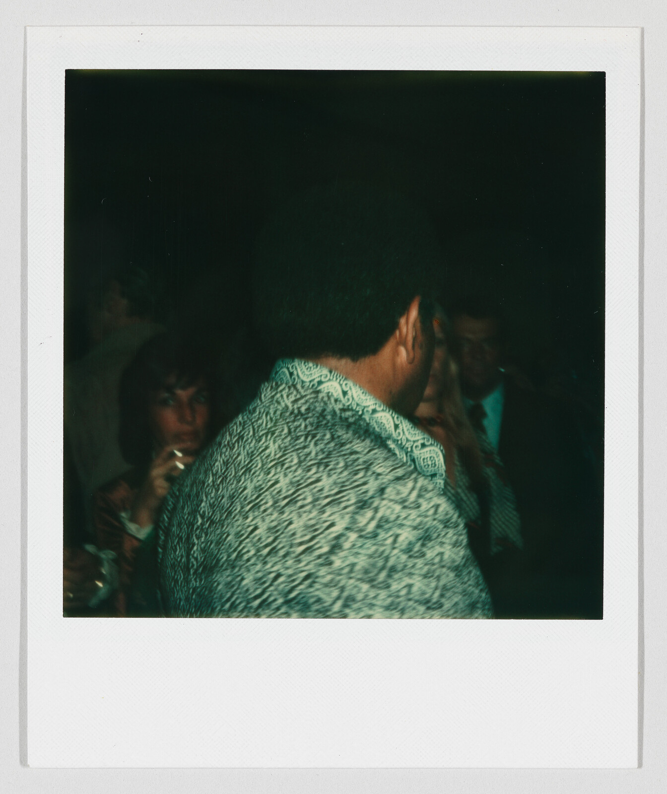 Man with patterned jacket seen from behind standing in a dim crowded room while others mingle.