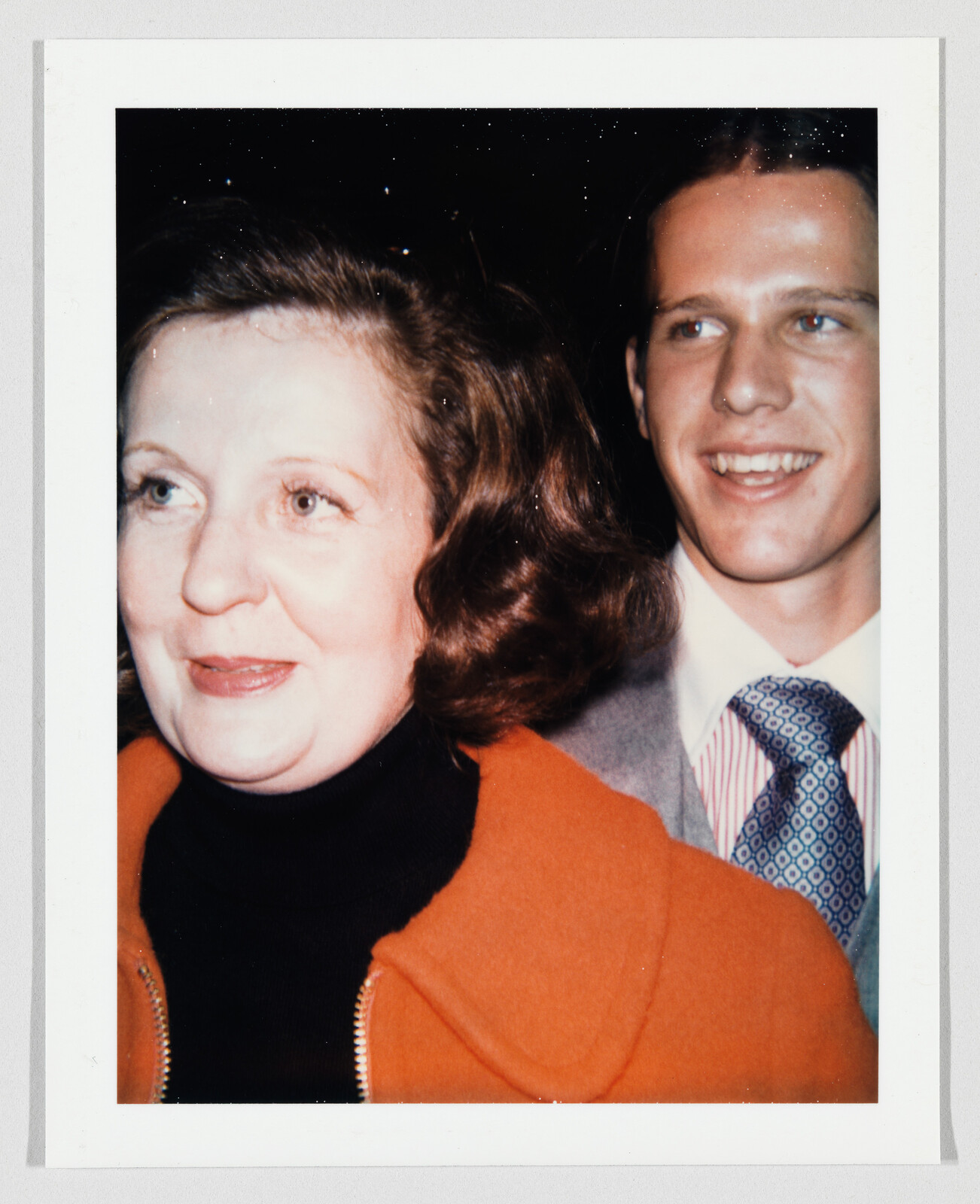 A woman in an orange coat stands in front of a smiling young man wearing a suit and tie.