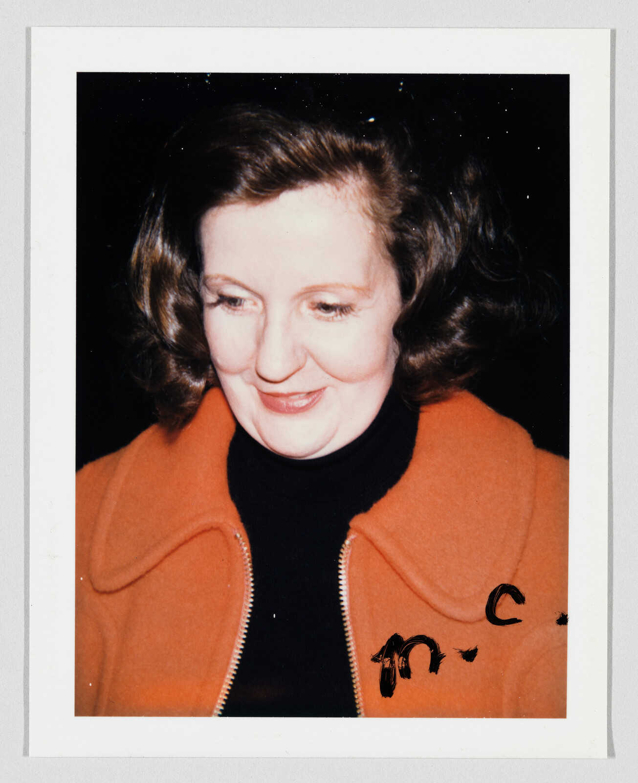 Woman with shoulder-length brown hair wearing an orange jacket and black shirt, looking down and smiling.