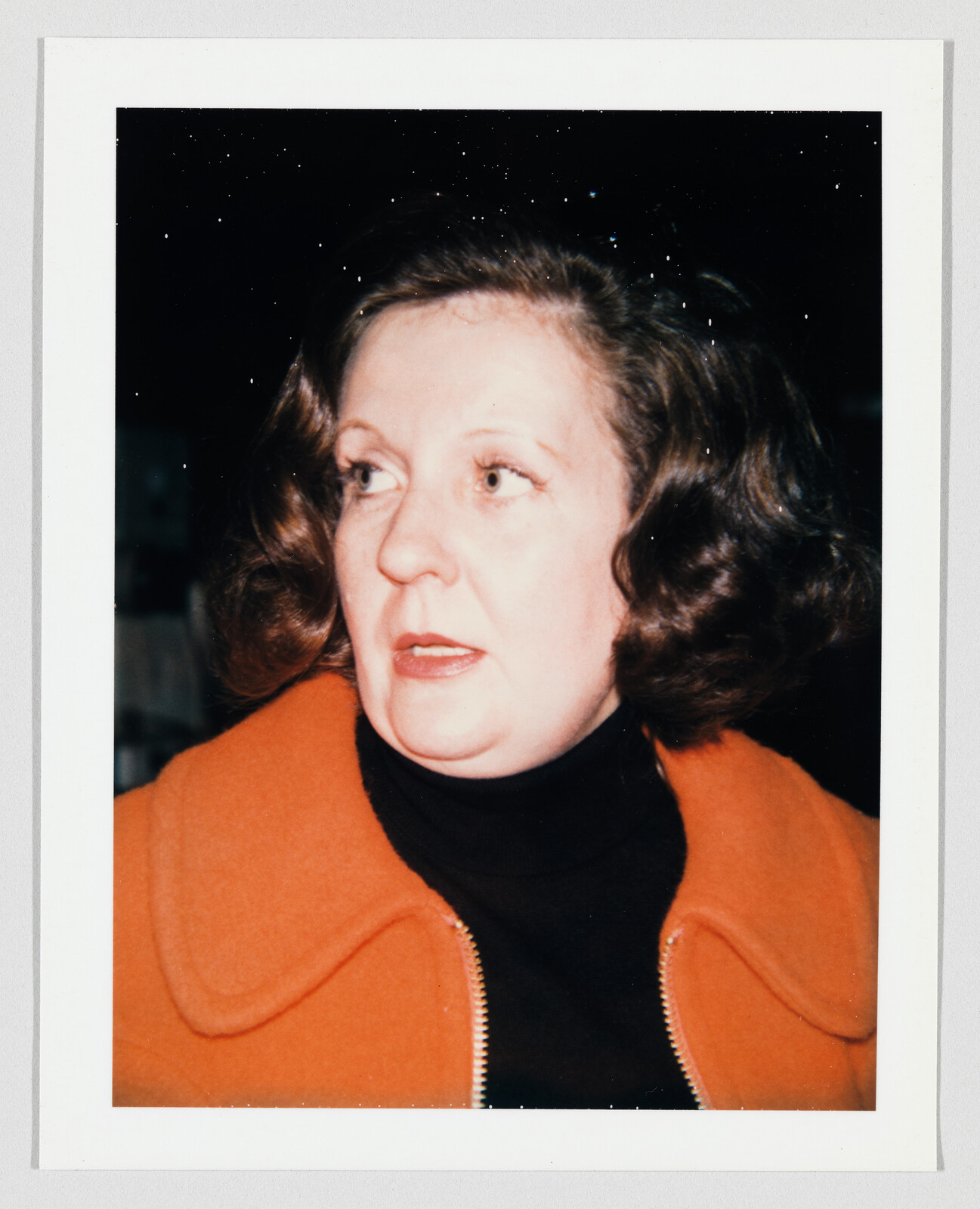 Woman with shoulder-length brown hair wearing an orange jacket and black turtleneck, looking to the side.