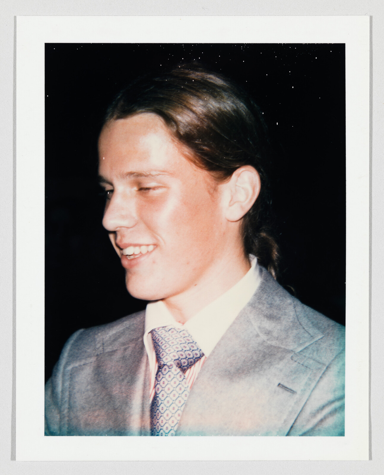 Young man with long hair tied back, wearing a light gray suit and patterned tie, smiling.