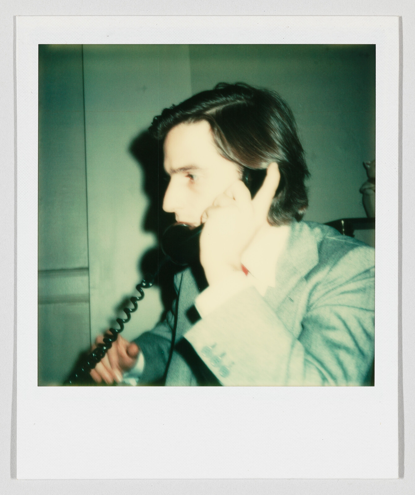 Young man in a suit talking urgently on a corded telephone inside a room.
