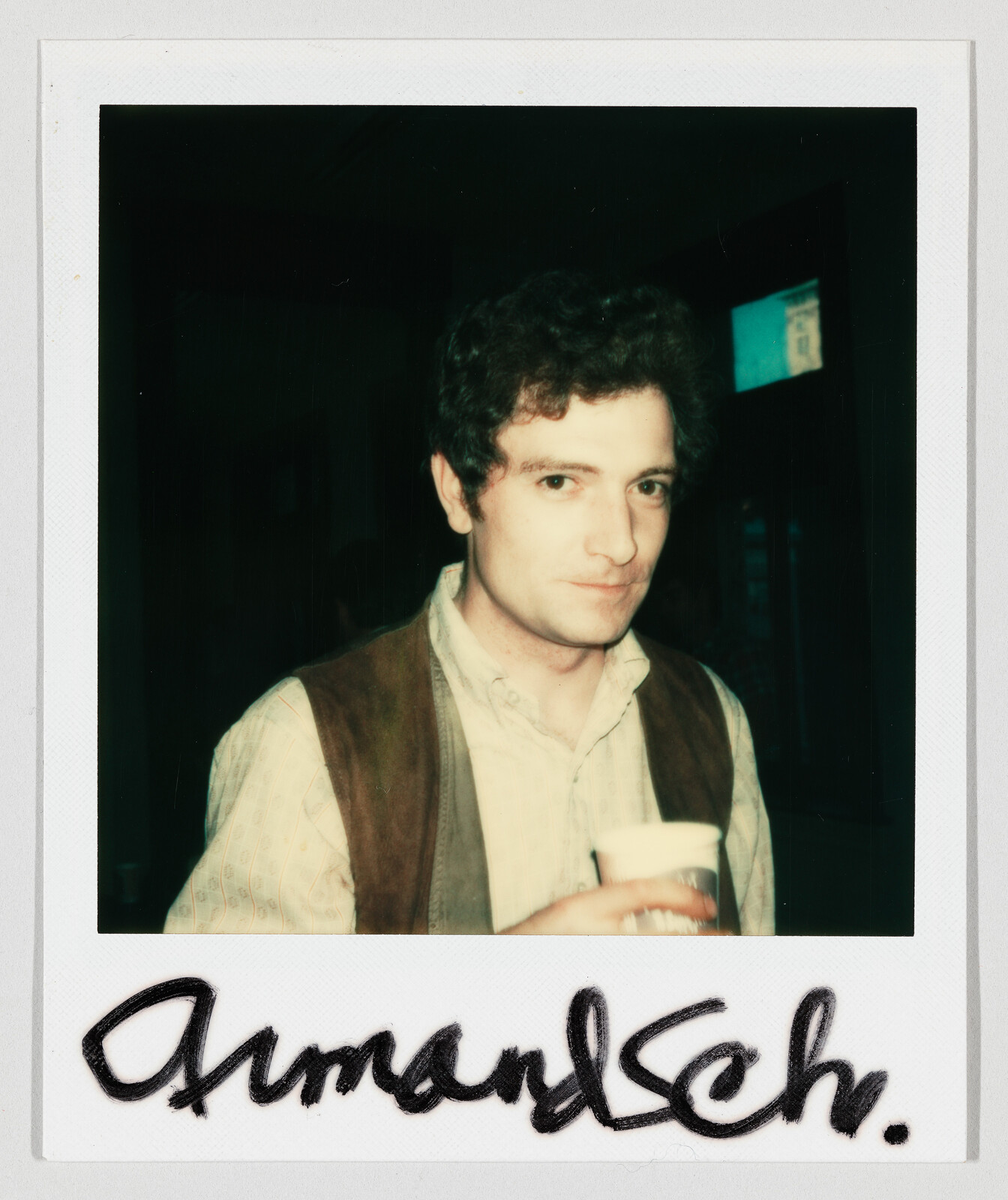 Young man with curly hair wearing a vest and holding a cup while looking at camera.