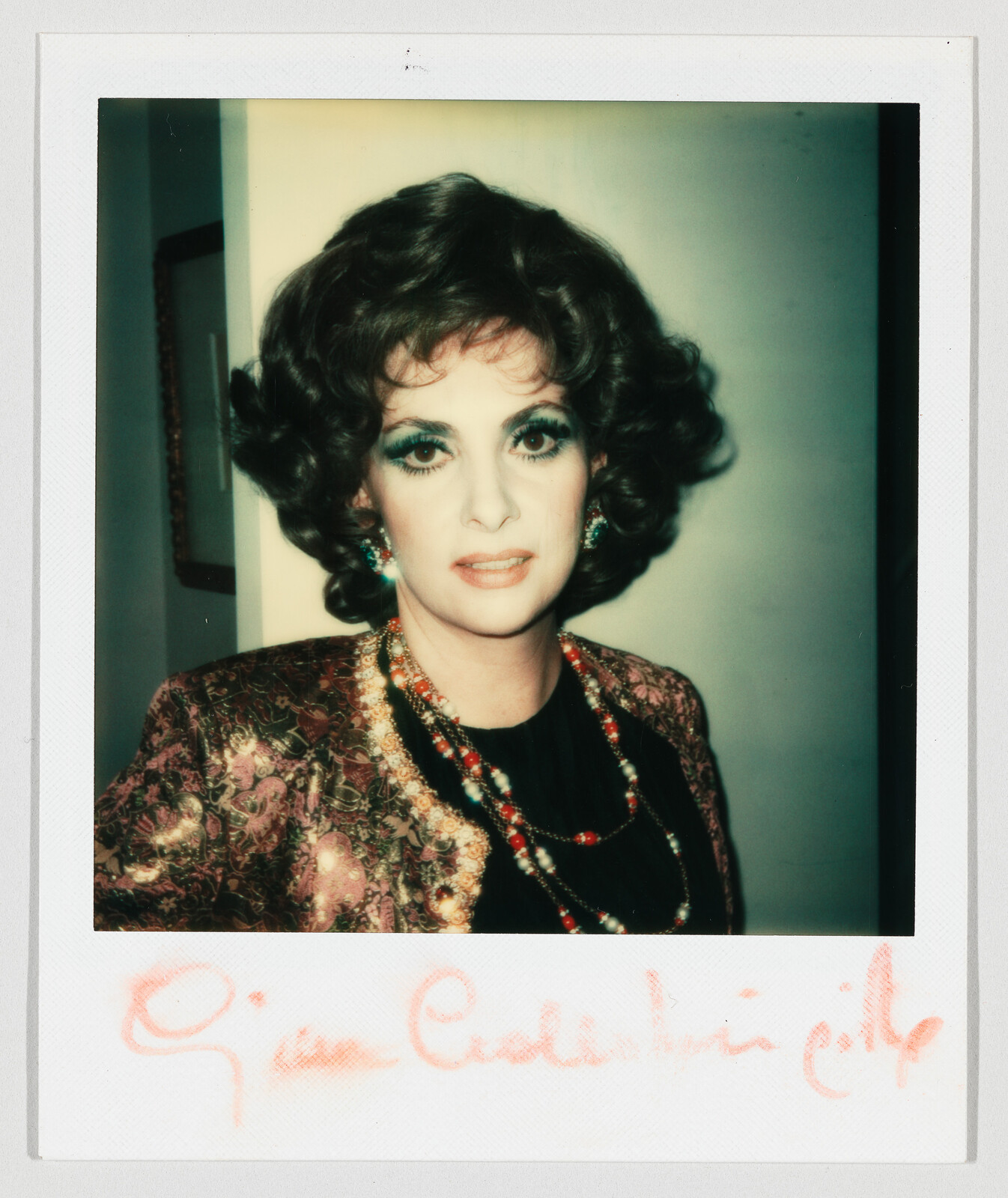 A woman with curled hair and heavy eye makeup wearing a patterned jacket and beaded necklaces.