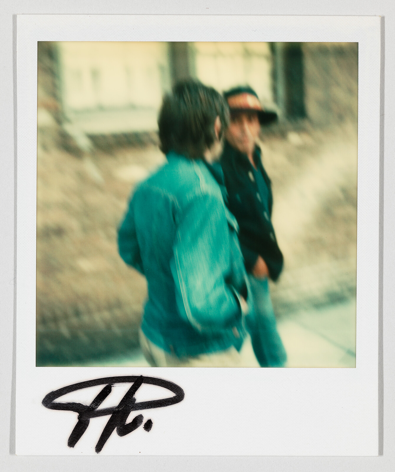 Two people walk and talk on a sidewalk, one wearing a denim jacket and the other a cap.