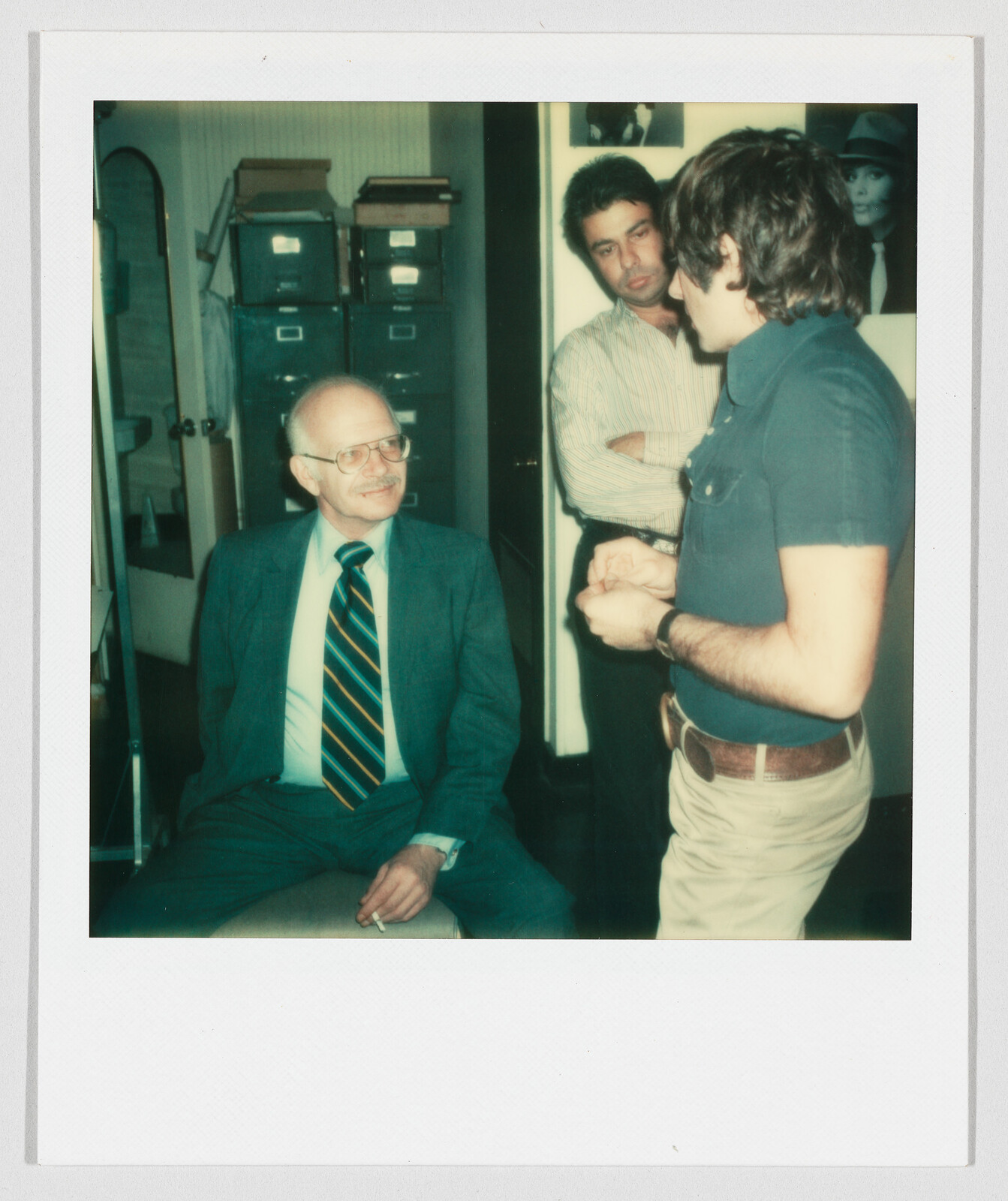 An older man in a suit sits smoking while two younger men stand talking in an office.