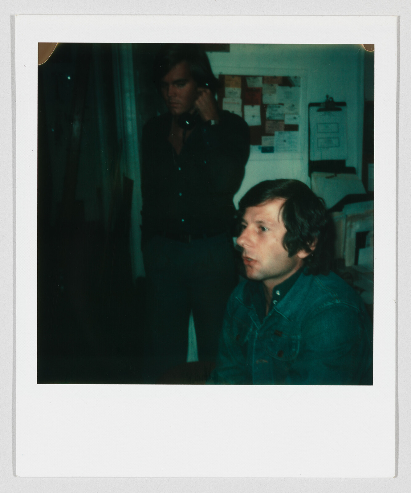 A man in a denim jacket sits looking to the right while another man talks on a phone.