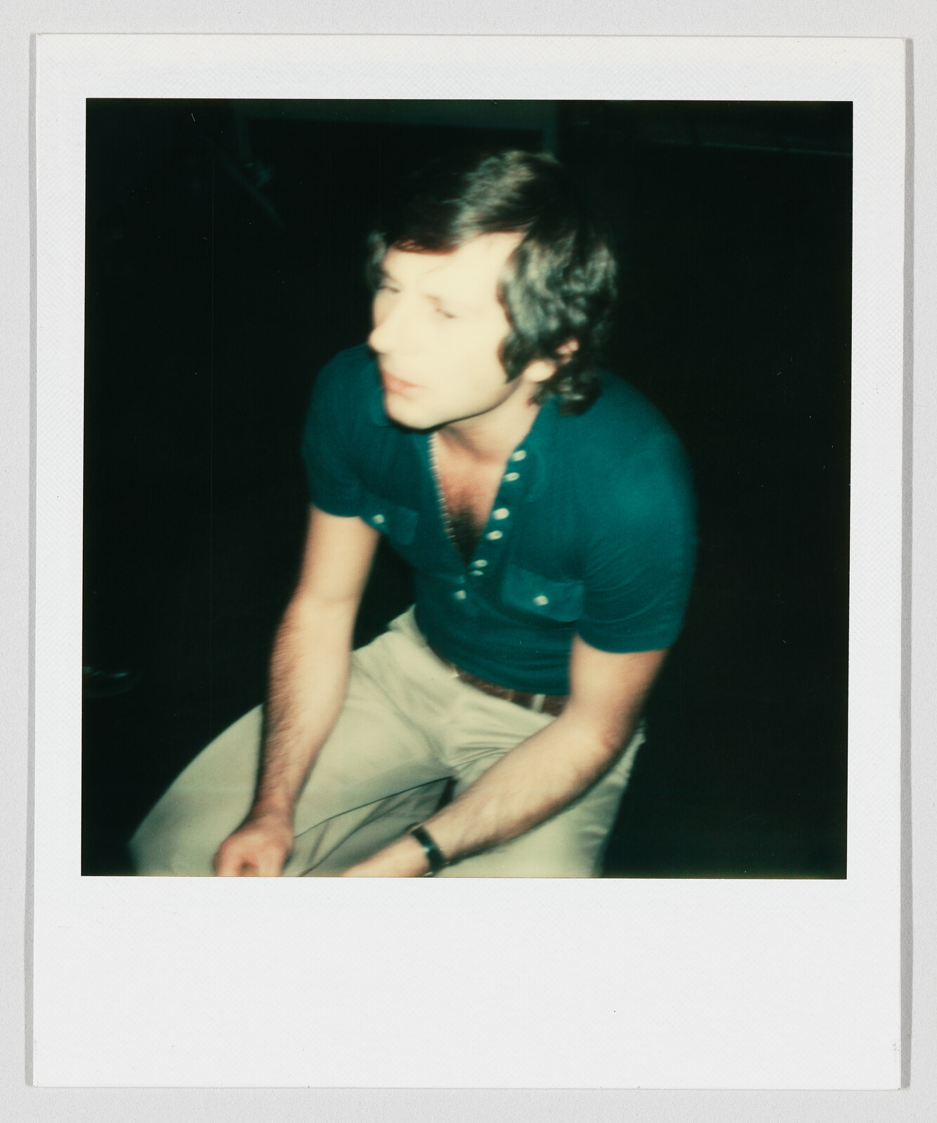 A young man in a teal shirt sits leaning forward with hands clasped, looking to the side.