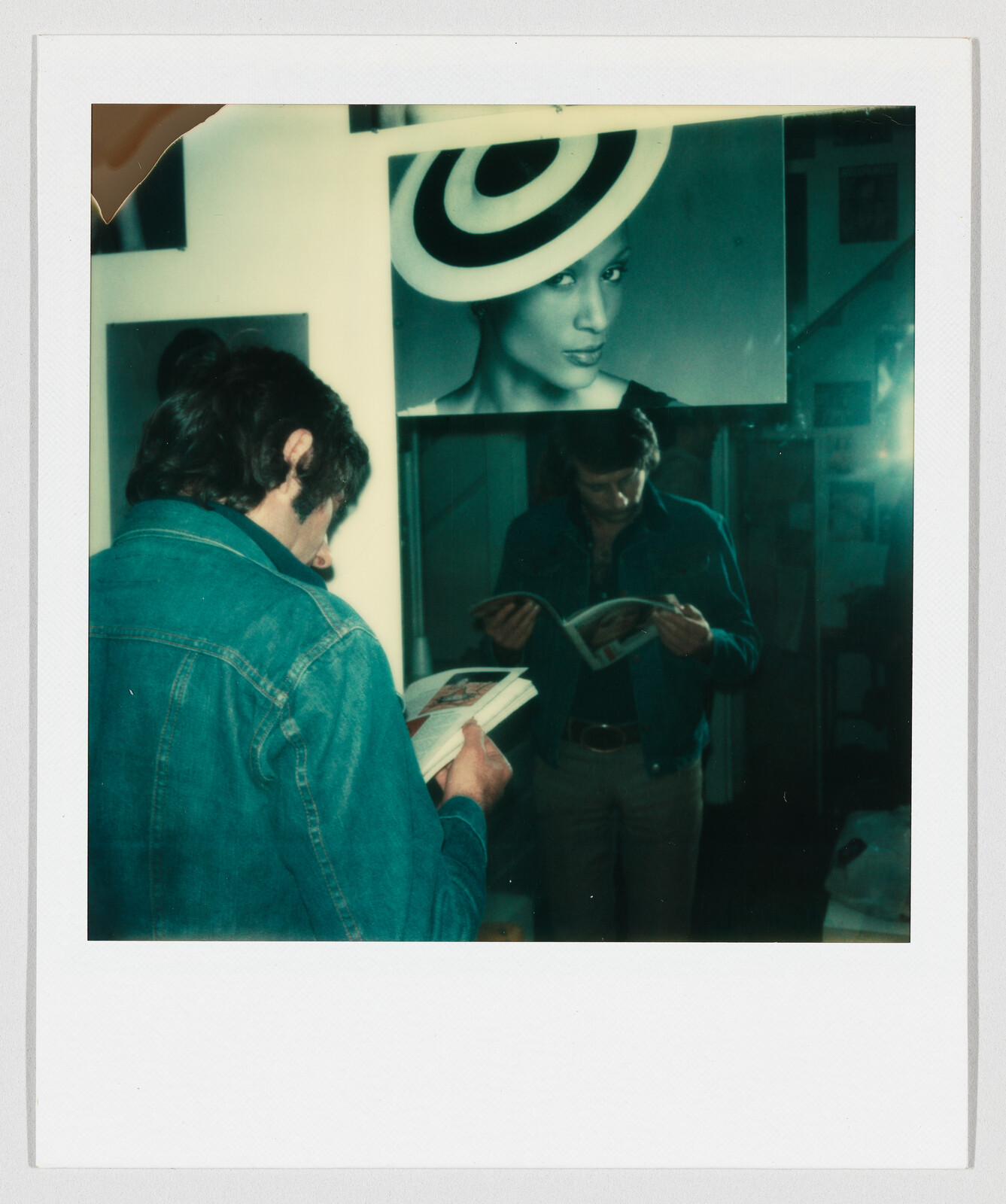 A man in a denim jacket stands reading a magazine while his reflection and a large portrait look on.